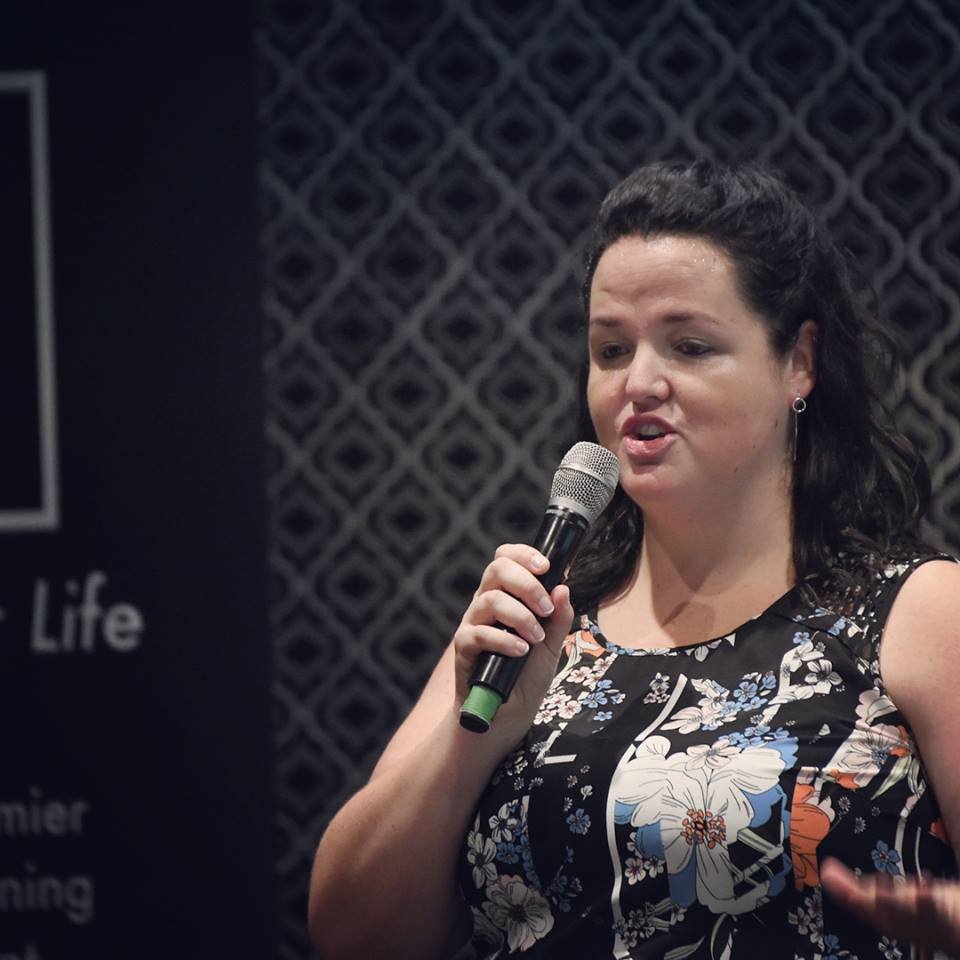 A woman with dark hair, wearing a black floral dress, speaking into a microphone at an indoor event with a patterned background.