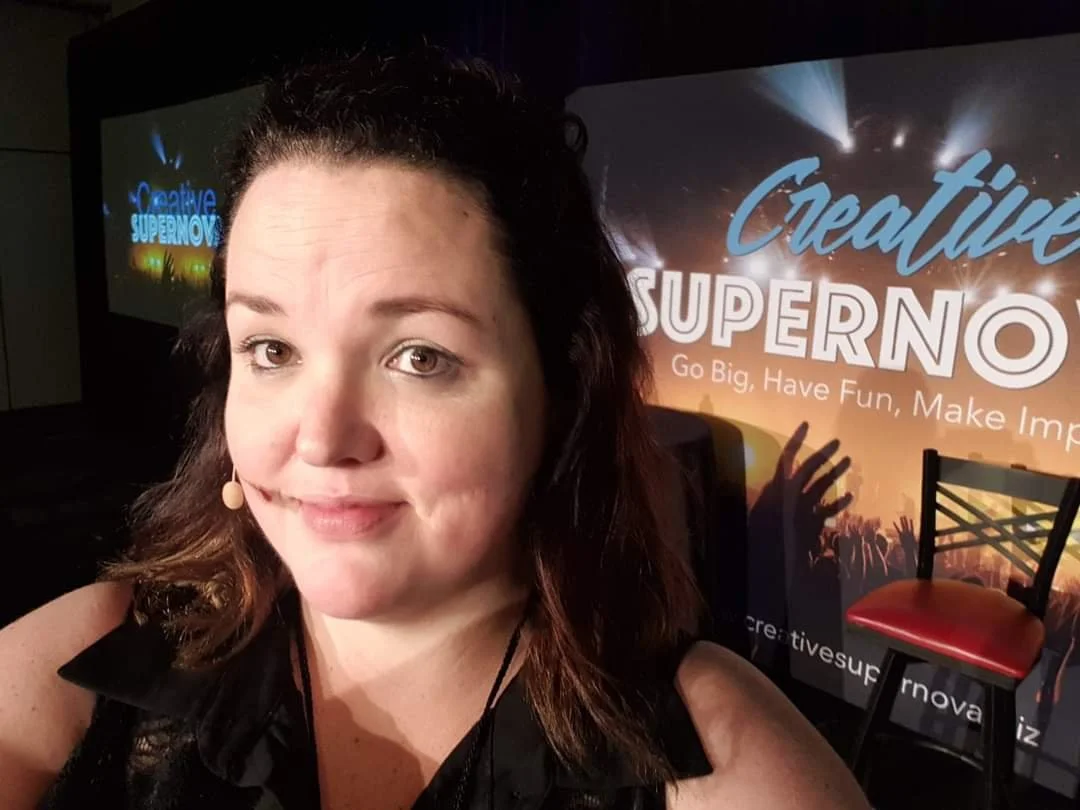 A woman taking a selfie at an event with a backdrop that reads "Creative Supernova" and "Go Big, Have Fun, Make Impact."