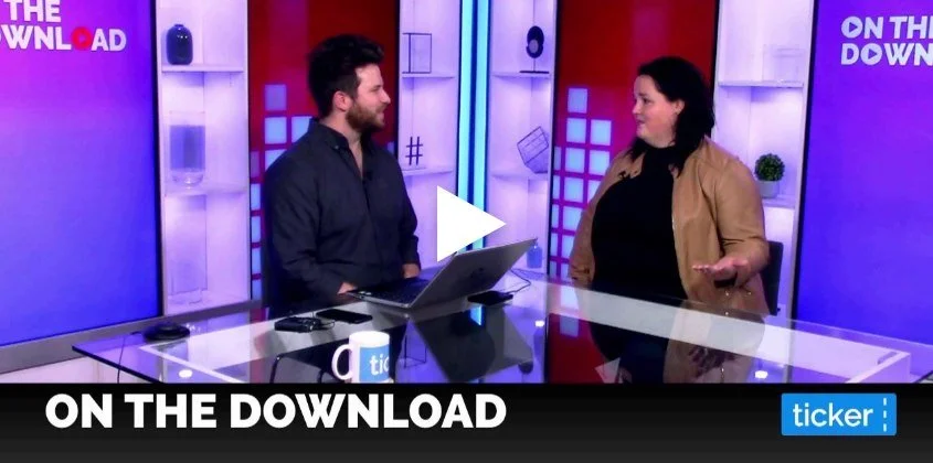 A man and a woman are having a conversation on a television set with a modern, colorful background. The man is standing behind a glass table with a laptop and a mug, and the woman is gesturing with her hand. The set has purple, blue, and red hues with graphics and text, including "ON THE DOWNLOAD" and "ticker."