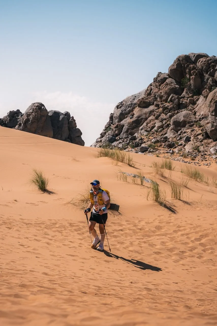 Hiker walking through a desert with sand dunes, sparse vegetation, and rocky formations.