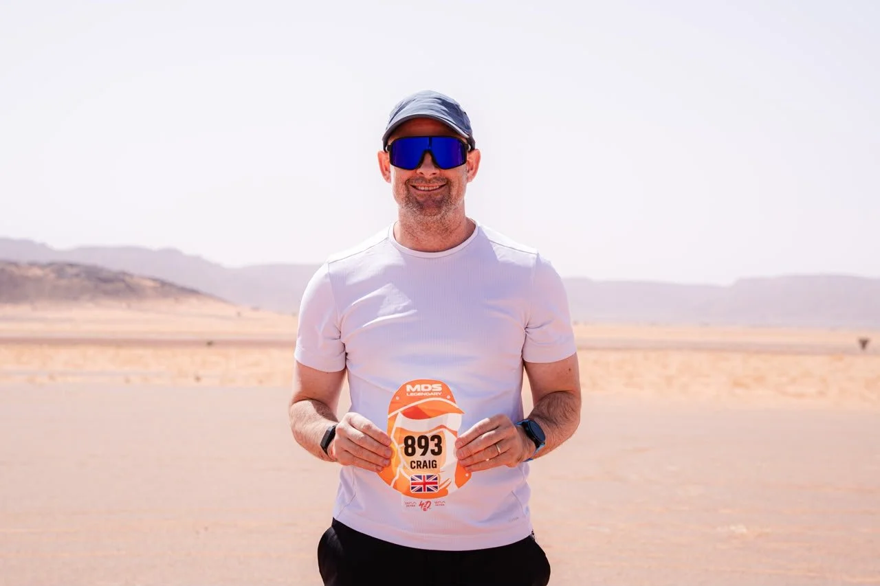 Man in white athletic shirt holding a race bib number 893 with the name Craig, standing in a desert landscape with mountains in the background.