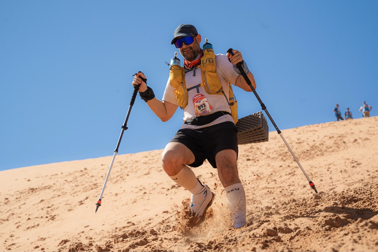 Man participating in a desert race, climbing sandy terrain with hiking poles, wearing a yellow hydration vest, sunglasses, and athletic gear.