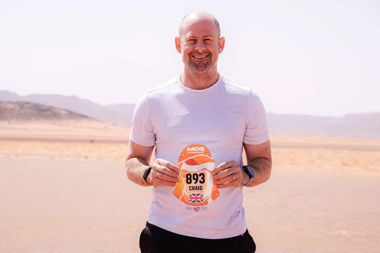 Man holding a race bib in a desert landscape, smiling, wearing a white T-shirt and a smartwatch.