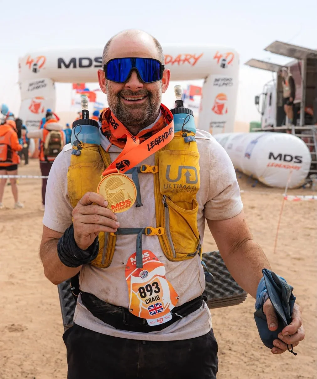 A man smiling and holding a gold medal after a race, wearing sunglasses, a yellow running vest, and a race bib with the number 893 and the name Craig. Behind him are other runners, an archway with the MDS logo, and an outdoor event setup.