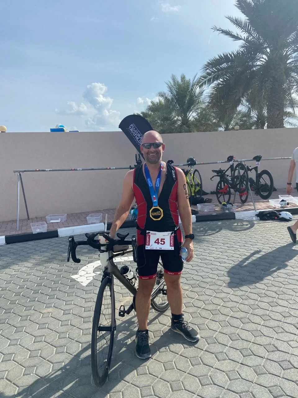 Smiling male triathlete with medal, standing next to his bicycle, at a sunny outdoor race event, with palm trees in the background.