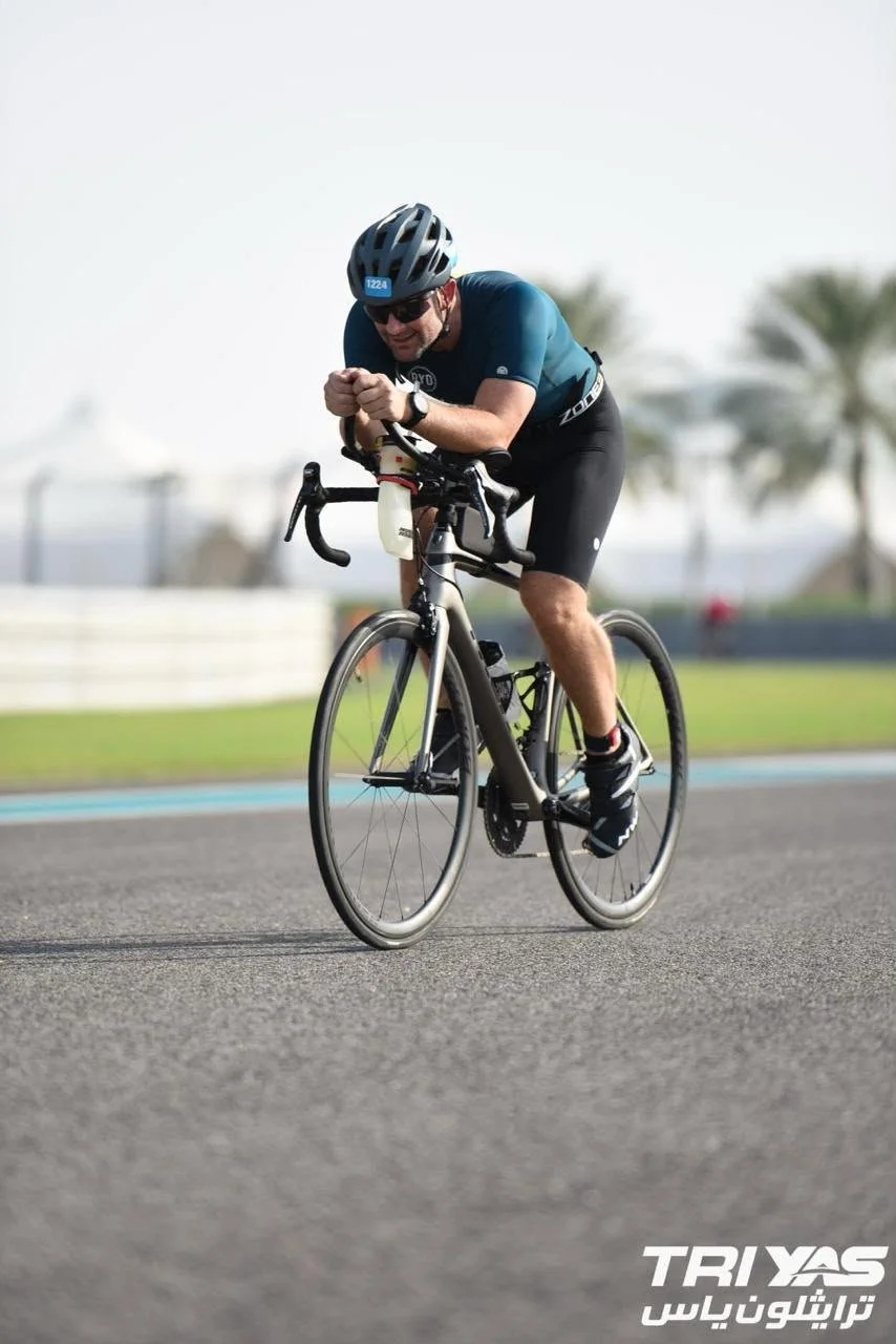 A man wearing a blue cycling jersey, black shorts, a helmet, and sunglasses riding a black road bike on a paved track with palm trees in the background.