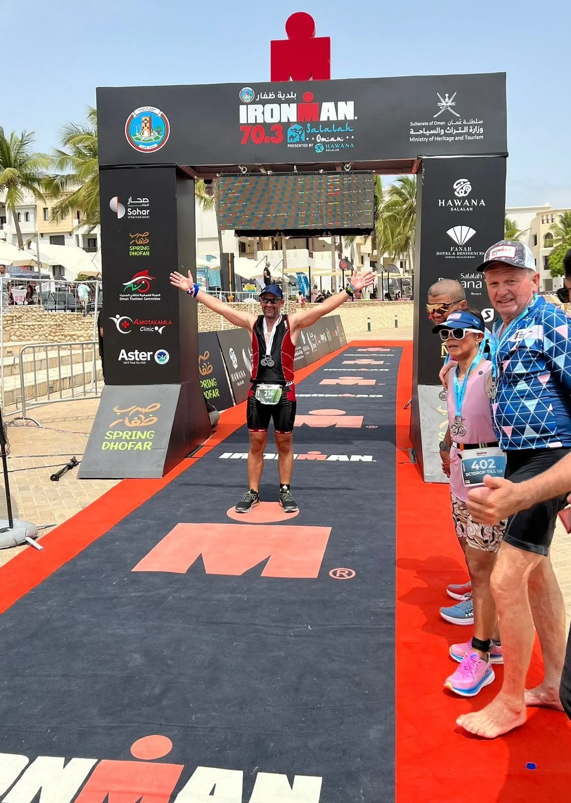 A man celebrating crossing the finish line of an Ironman race at the Spring Dhofar event in Salalah, Oman, with supporters nearby, palm trees in the background, and a black finish line arch with event logos.