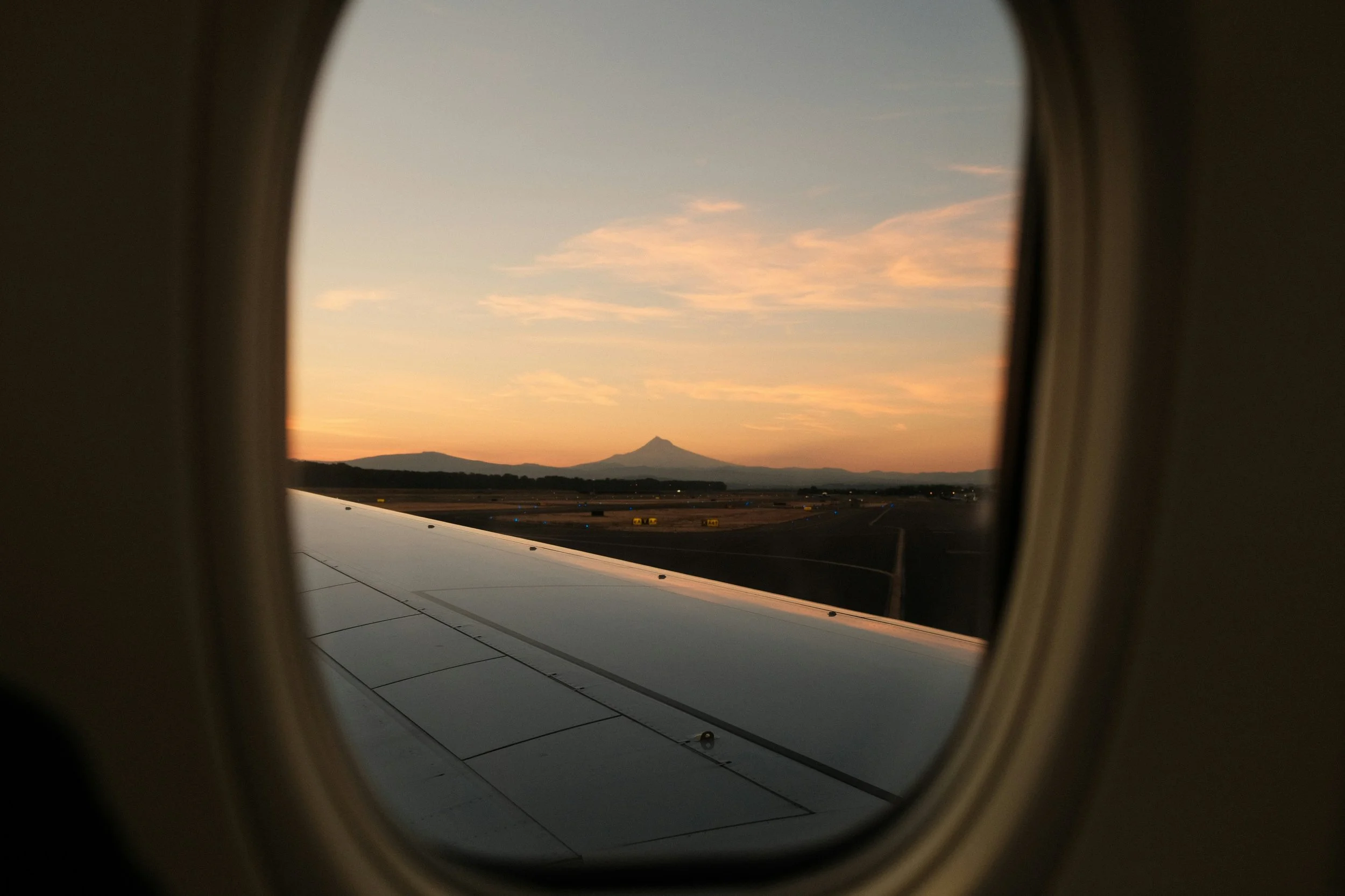 View of a mountain, likely Mount Fuji, seen through an airplane window during sunset or sunrise with light orange and pink clouds in the sky.