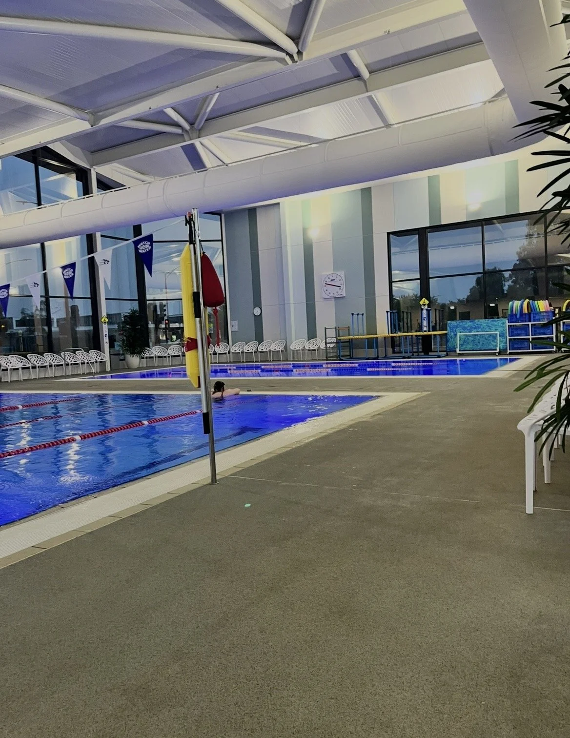 Indoor swimming pool with lounge chairs, a swimmer in the water, and pool safety equipment; large windows and a clock on the wall.
