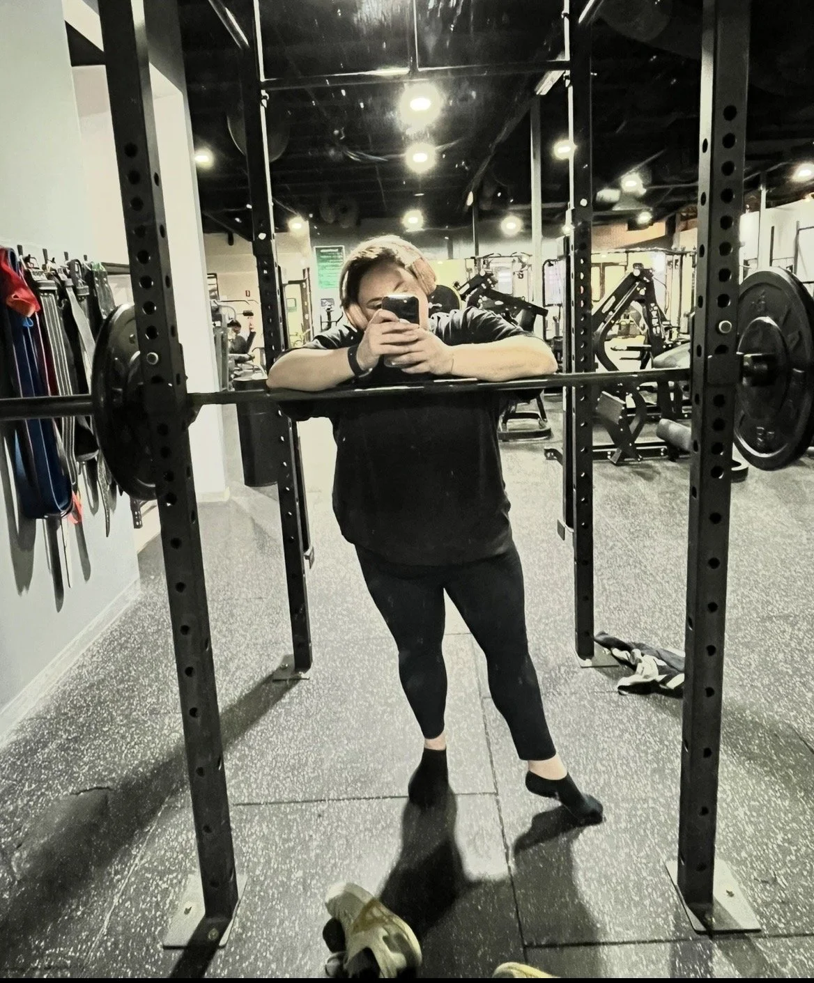 A person taking a mirror selfie at the gym, standing in front of a squat rack with weight plates and gym equipment in the background.