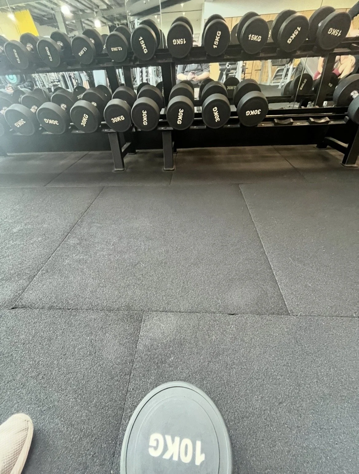 View of a gym with a row of black dumbbells with weights labeled from 15 kg to 30 kg on a rack, a mirror reflecting people and gym equipment, and a black rubber gym floor. The photo appears to be taken by someone standing or sitting on the floor, with part of their shoe visible at the bottom of the image.
