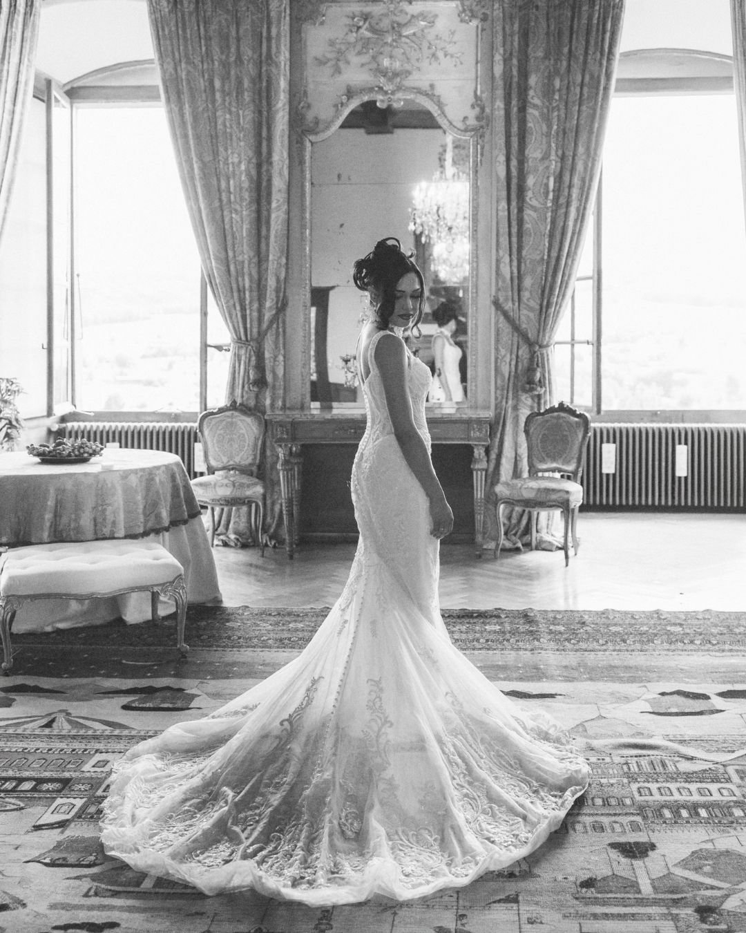 Une femme en robe de mariée dans une pièce élégante avec un grand miroir, deux fauteuils et une table avec un plateau de fruits, en noir et blanc.