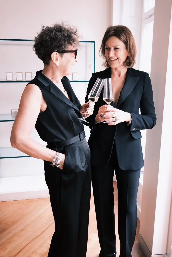 Deux femmes d'âge mûr, habillées en noir, souriant tout en se tenant par des verres de champagne dans une ambiance élégante.