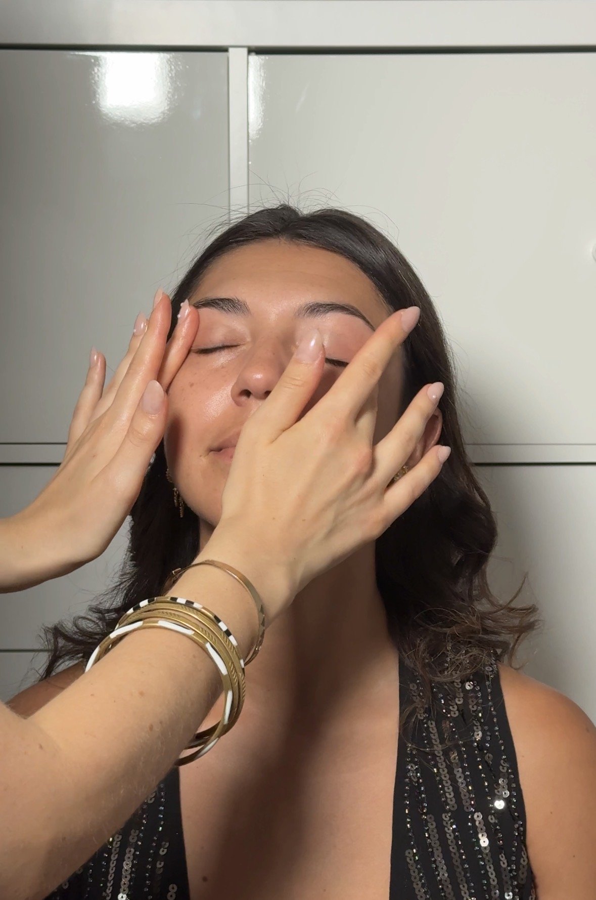 Une femme en robe noire à sequins reçoit un massage du visage avec ses yeux fermés, par des mains portant des bracelets dorés et une manucure claire, devant un mur blanc.