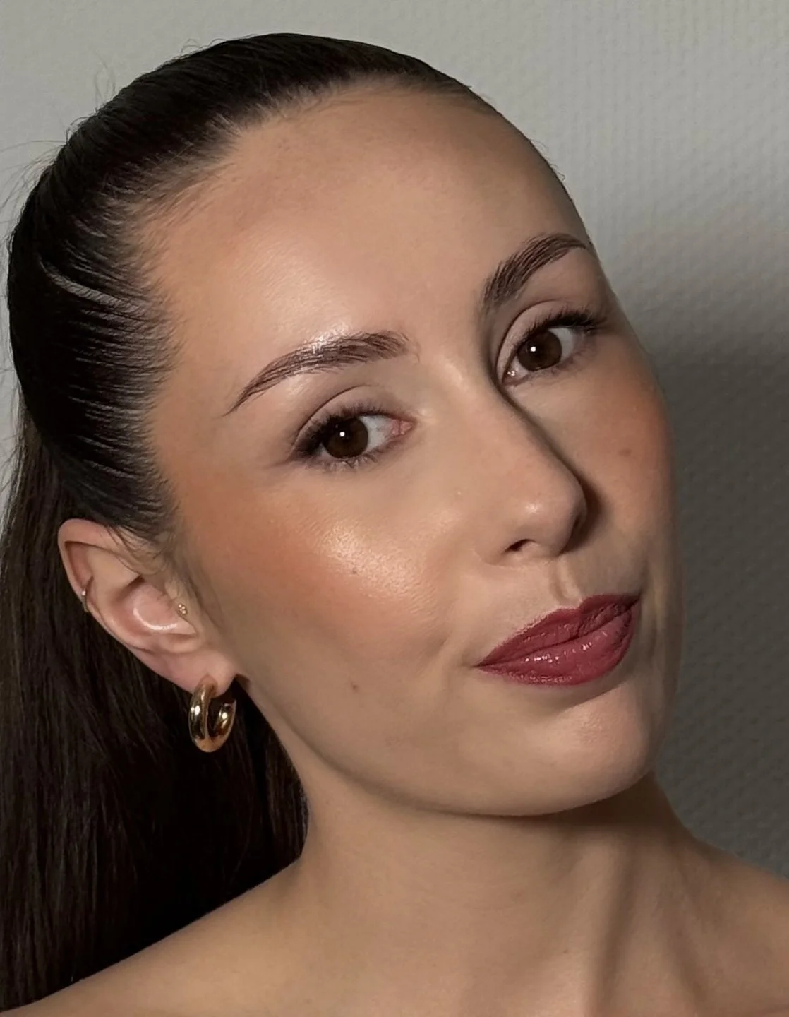Une femme avec des cheveux bruns lissés, portant des boucles d'oreilles en or, maquillée avec des lèvres rouge foncé et un maquillage naturel.