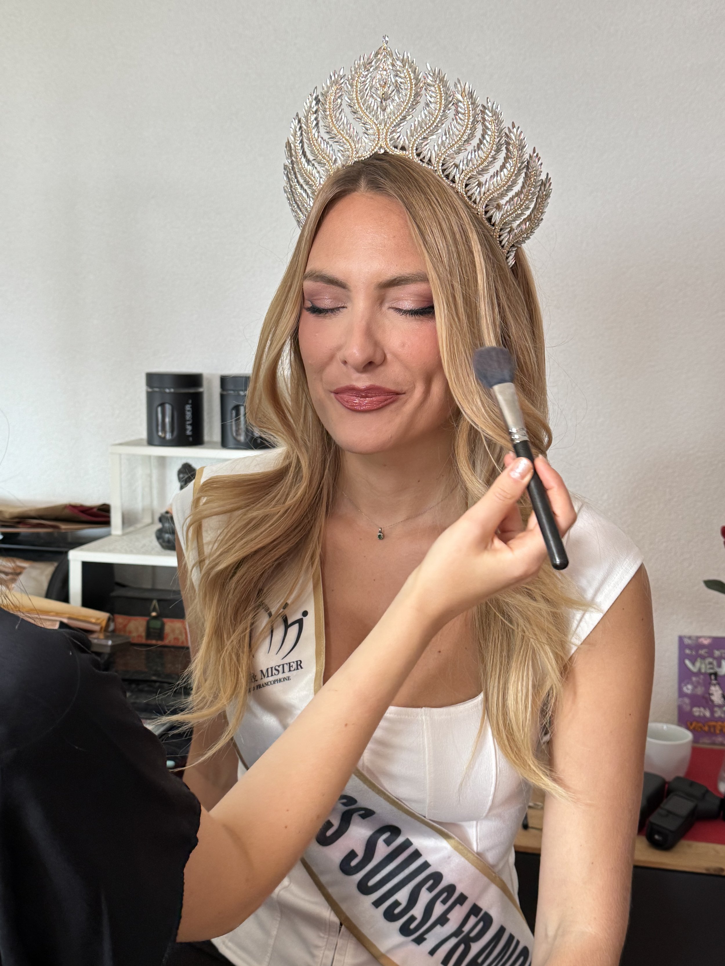 Une femme portant une couronne brillante et un sash participe à un concours de beauté, un maquilleur applique du maquillage sur son visage.