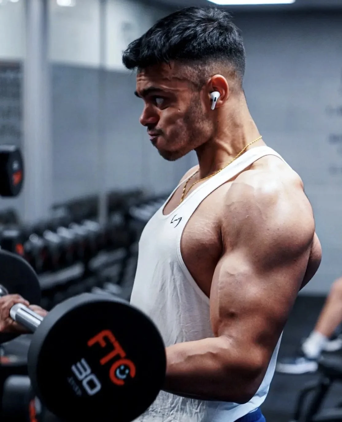 A young man with a muscular build lifting a dumbbell in a gym, wearing a white tank top, wireless earbuds, and a gold chain, focused on his workout.