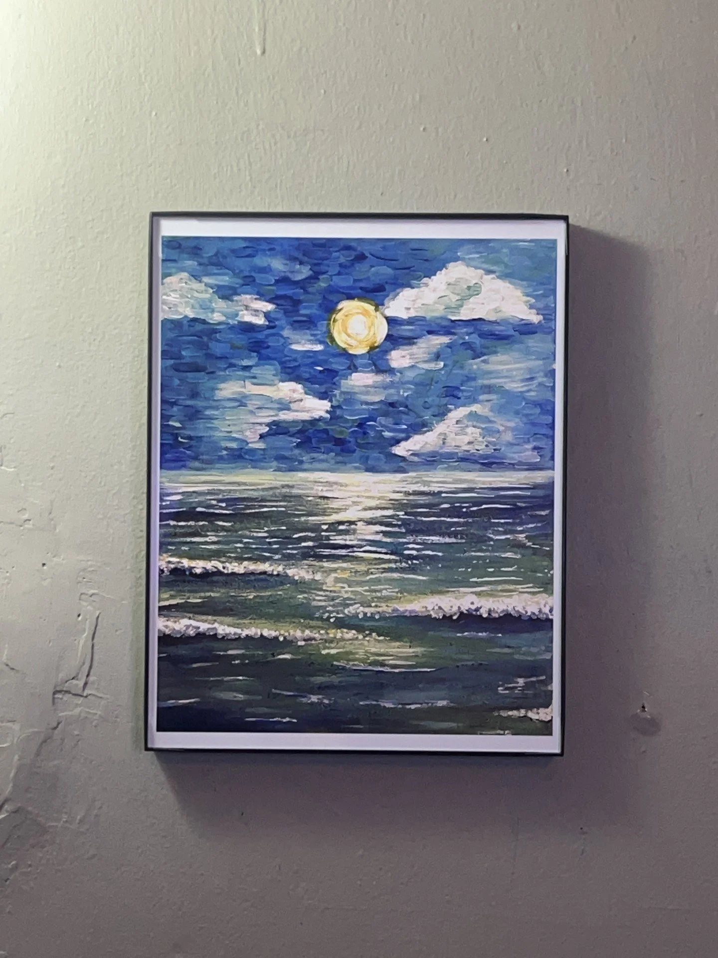 A framed painting of a seascape with a full moon, clouds, and waves, hanging on a textured wall.