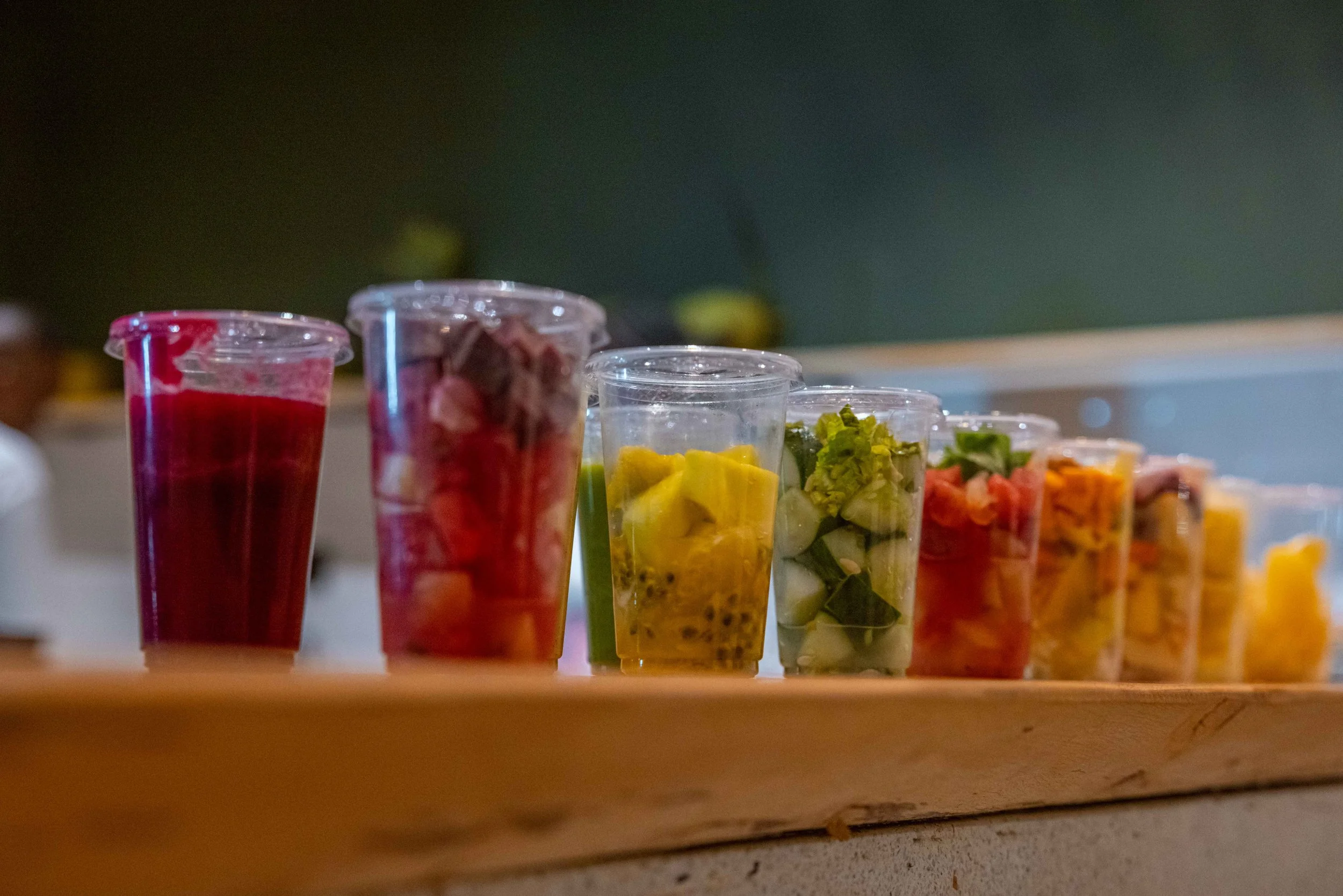 Rows of clear plastic cups filled with colorful chopped fruits and vegetables, lined up on a wooden surface.