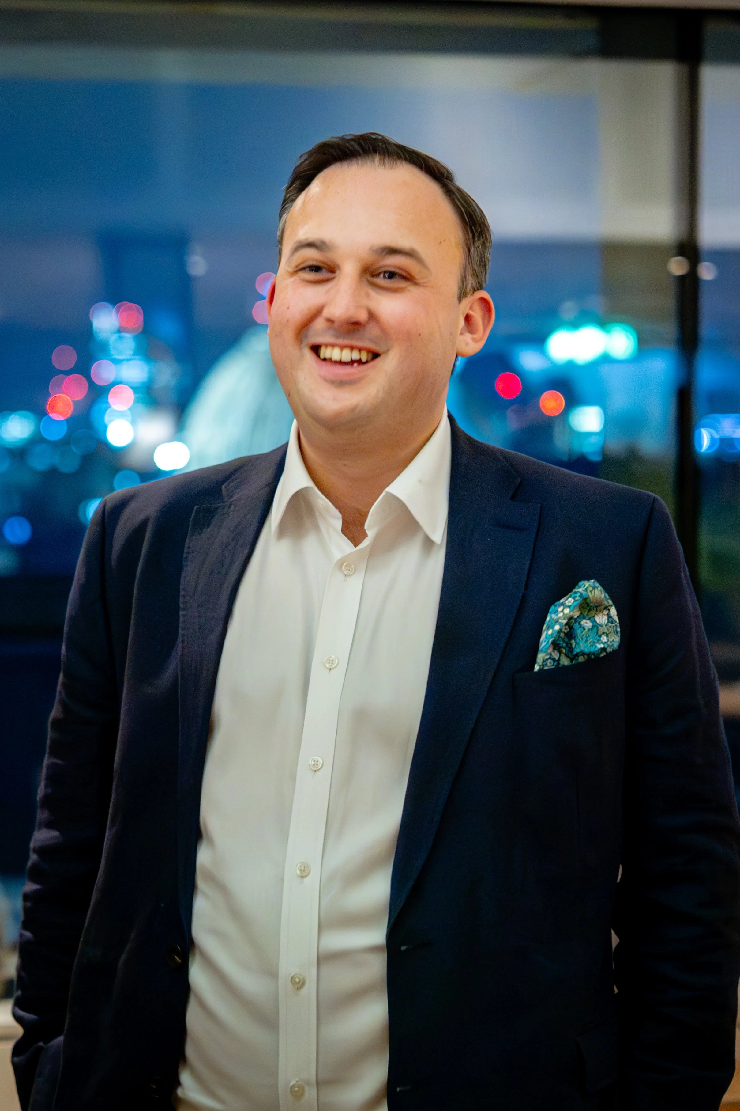 A smiling man in a business suit with a white dress shirt and a patterned pocket square, standing inside with a cityscape view at night in the background.