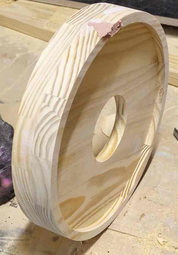 A round piece of light-colored wood with a central hole, standing upright on a workbench. The wood has visible grain patterns and some rough, unpolished edges.