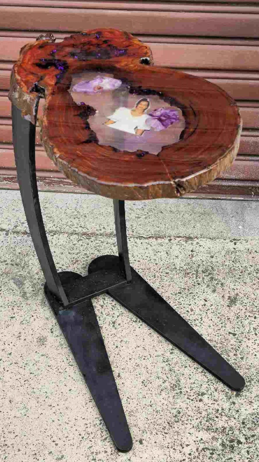 A wooden side table with a band-sawed cross-section top featuring a picture of a woman in a white shirt inside a resin inlay, placed on a black stand against a brown wooden wall background.