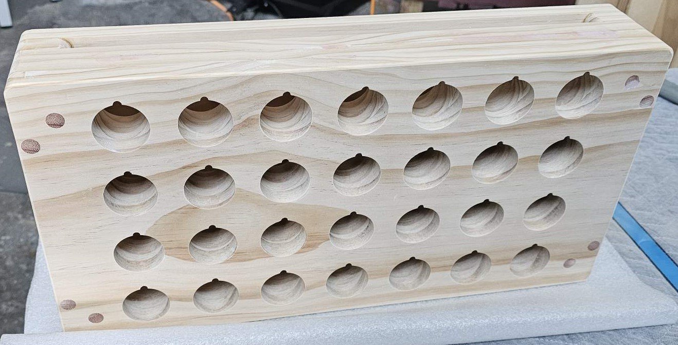 Unfinished wooden wine rack with 25 circular holes for bottles, placed on a white surface.