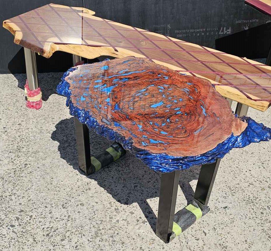 Two wood tables with unique designs; one has a smooth, natural wood finish with a protective coating, and the other has a textured, painted, and carved surface resembling a swirling pattern with blue and red colors.