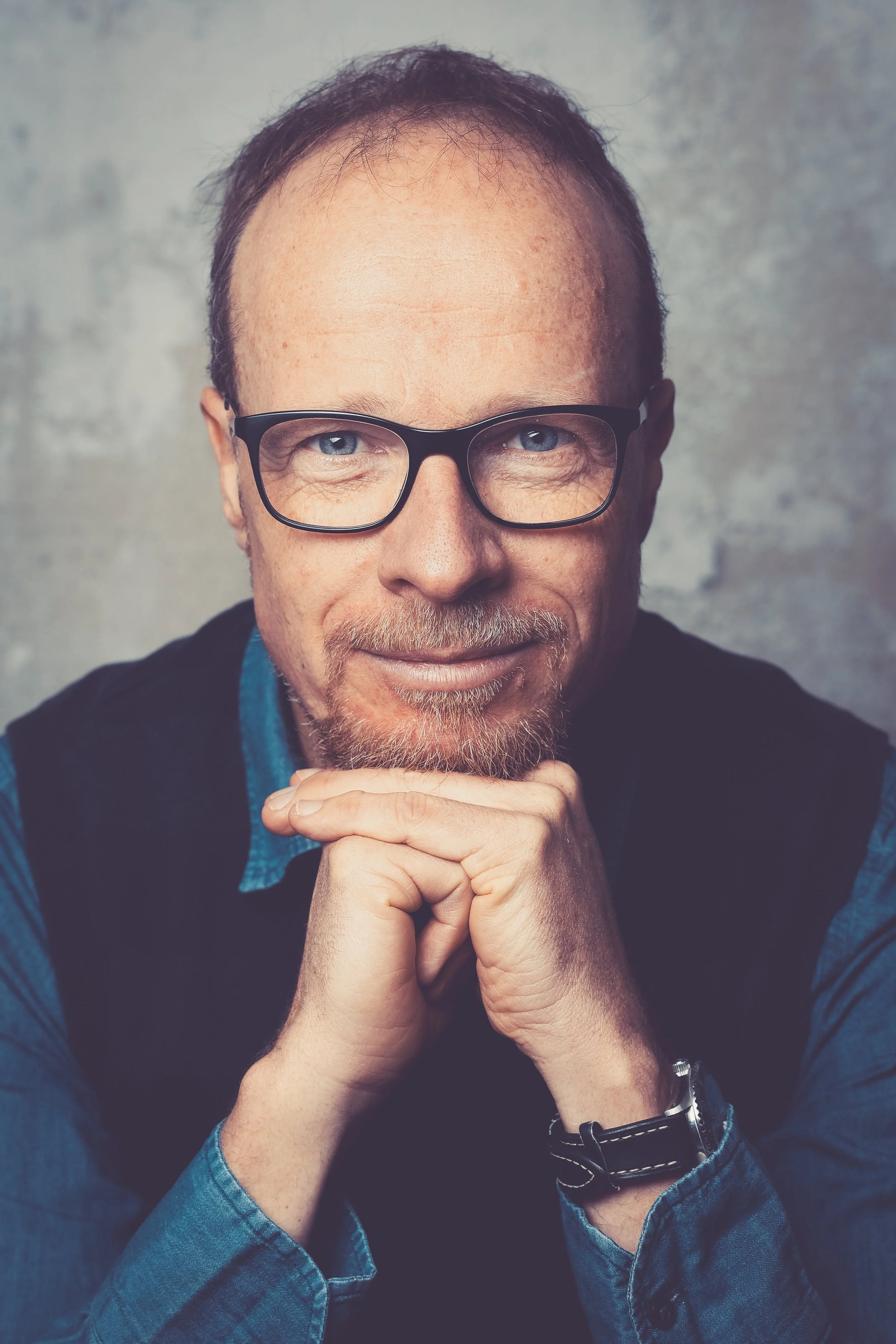 Porträt eines lächelnden Mannes mit Brille, kurzen roten Haaren, Bart und Brille, der die Hände unter seinem Kinn verschränkt und auf die Kamera schaut.