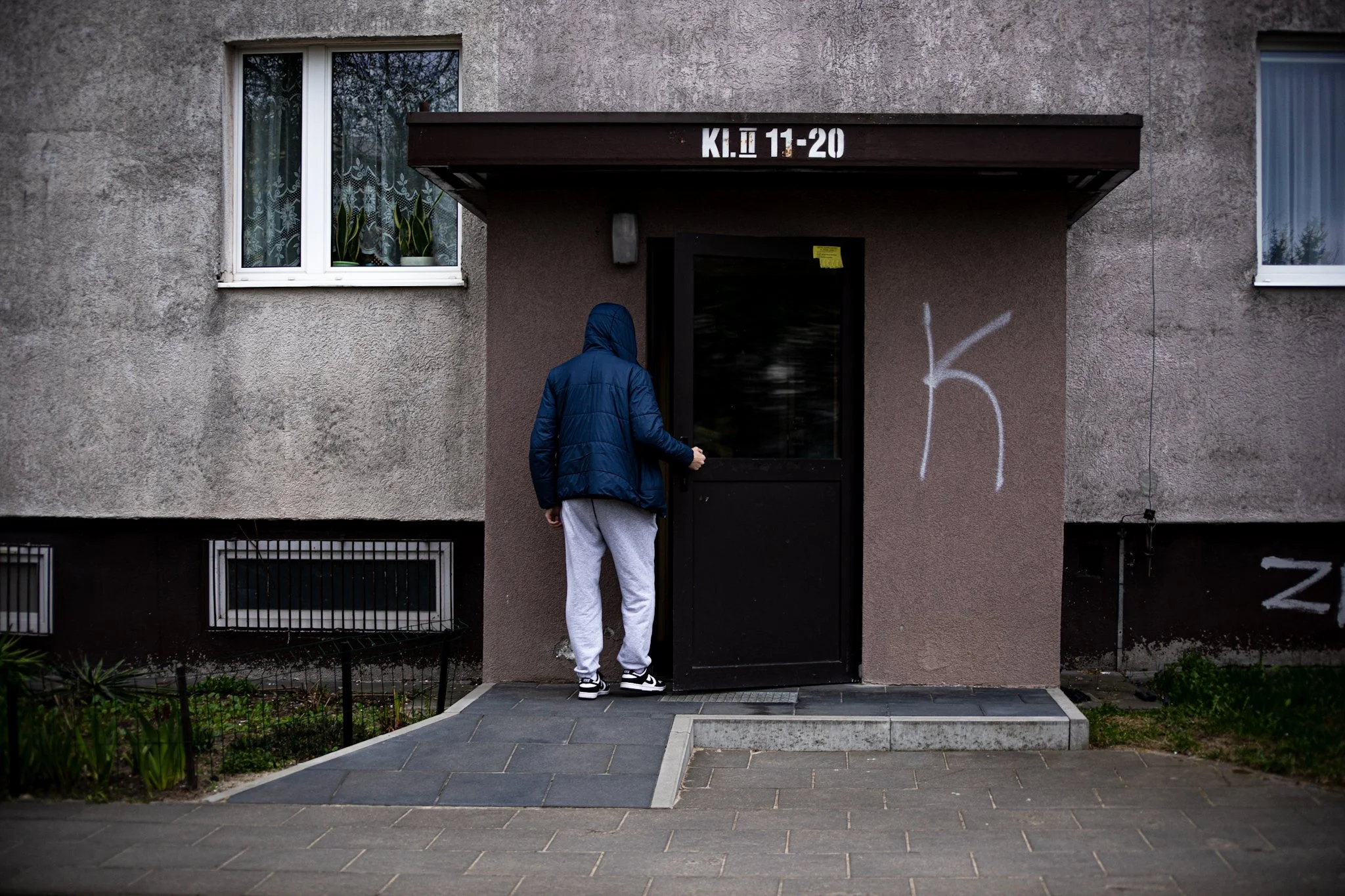 Person in a blue jacket and gray sweatpants standing outside a gray concrete apartment building door, with graffiti on the wall and potted plants in the windows.