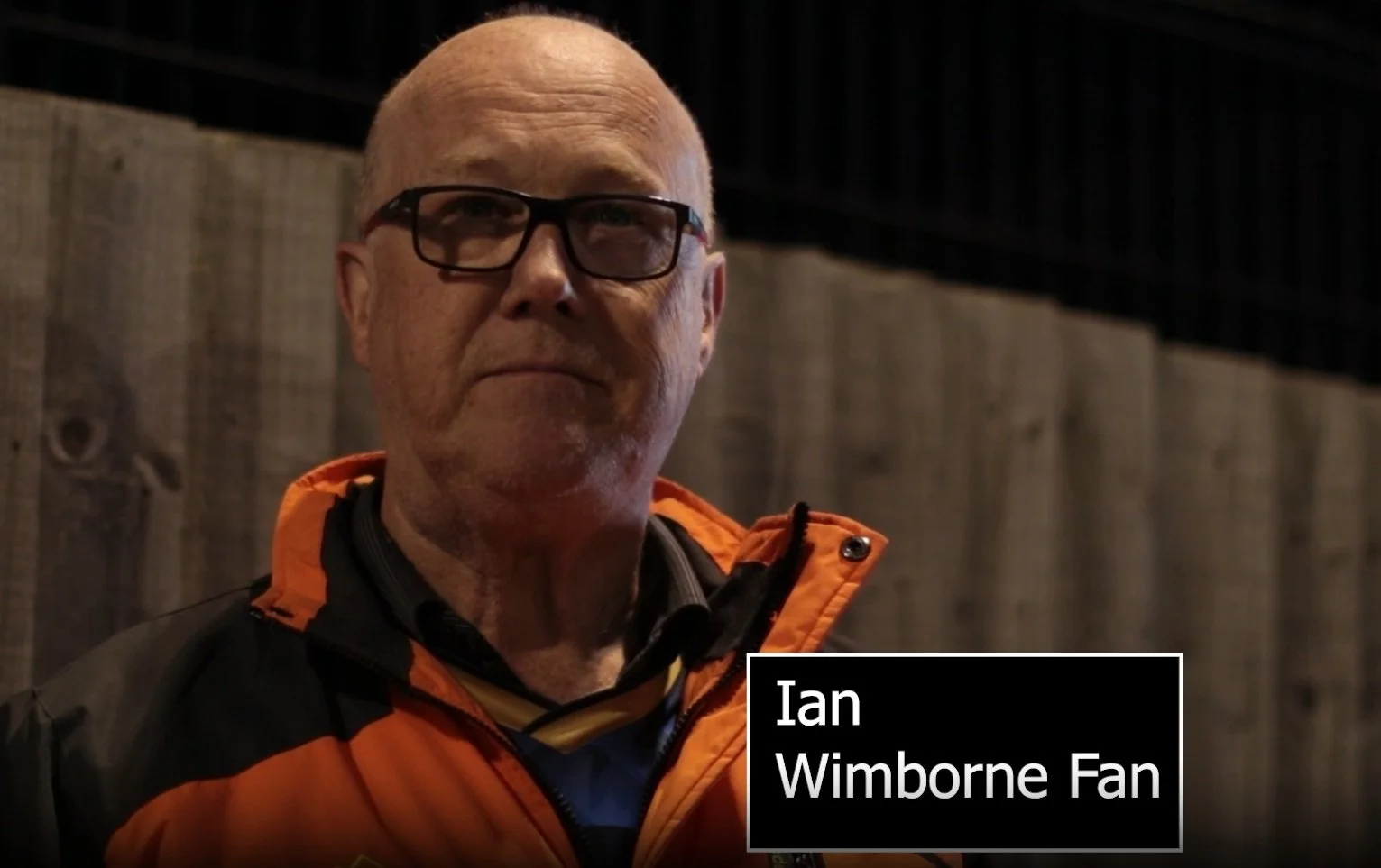 A middle-aged man with glasses and a bald head wearing an orange and black jacket, standing outdoors at night in front of a wooden fence.