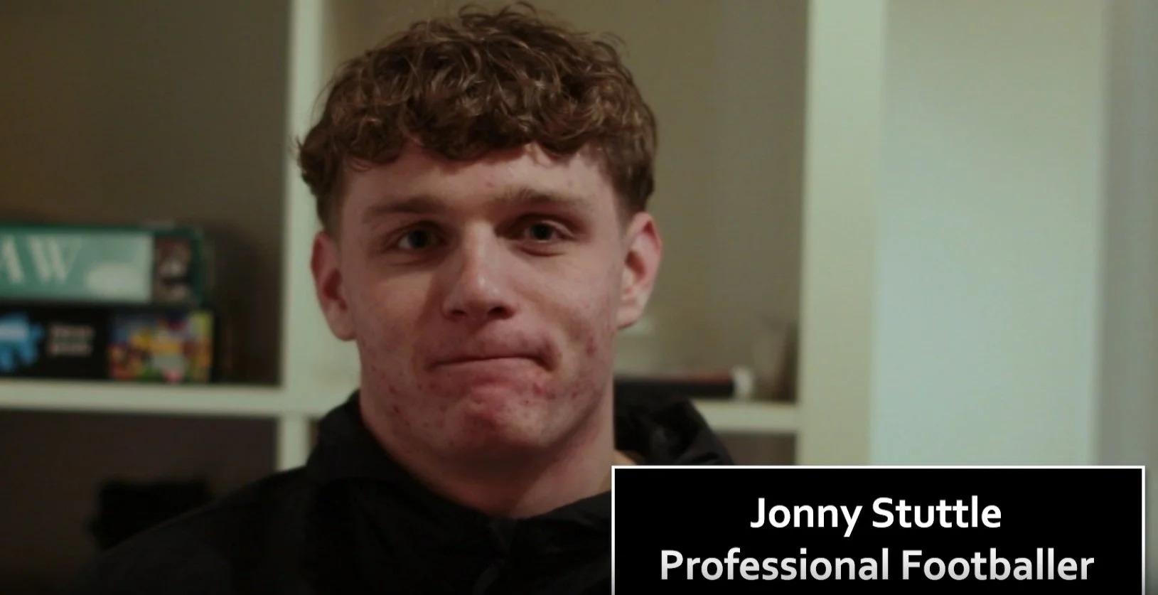 A young man with short curly hair and acne, wearing a black hoodie, in a room with shelves in the background. The caption reads: 'Jonny Stuttle, Professional Footballer.'