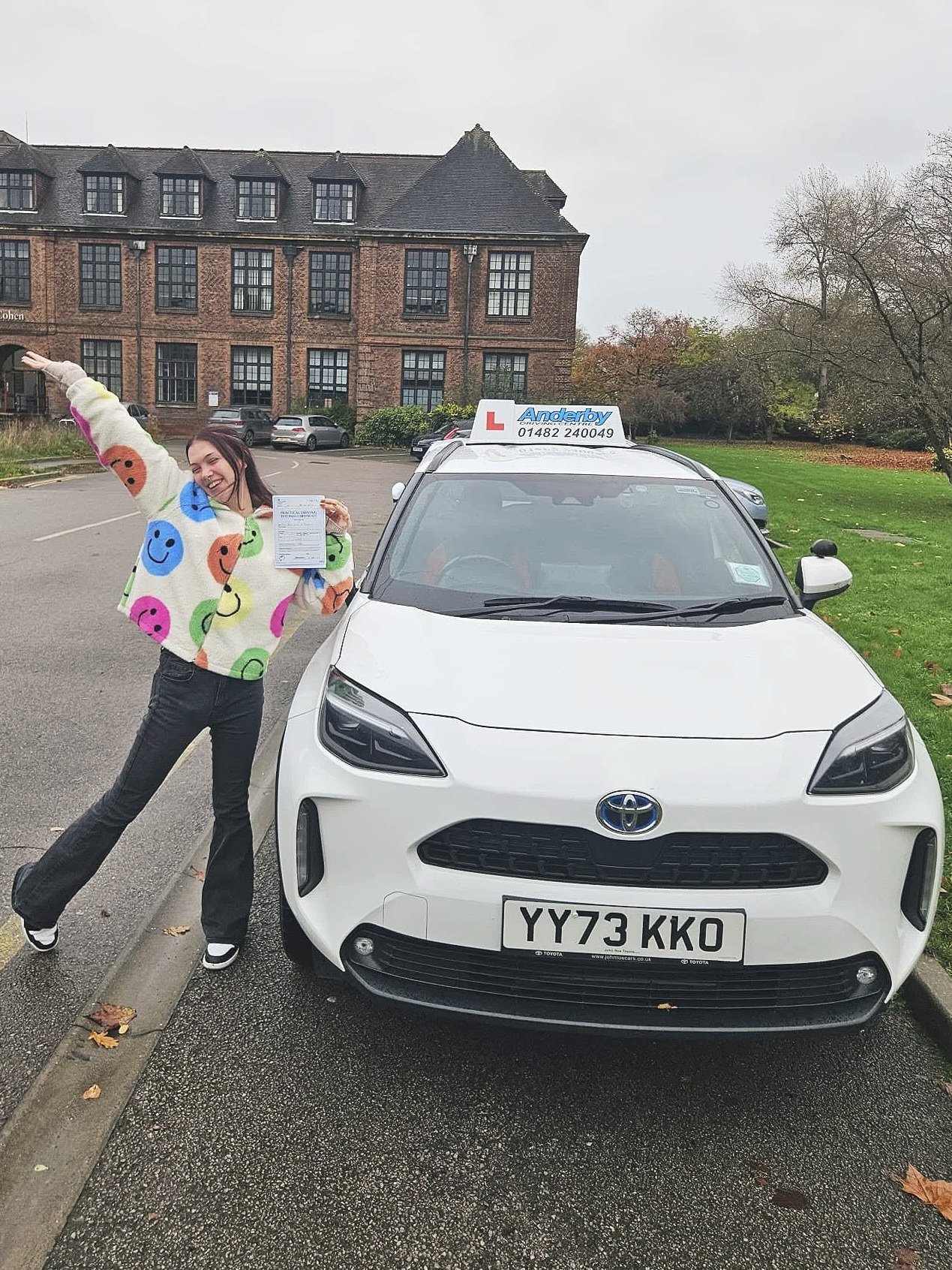 A woman wearing a colorful smiley face sweater is standing next to a white Toyota car with a driving school sign on top, in an outdoor parking lot on a cloudy day, holding a document and smiling.