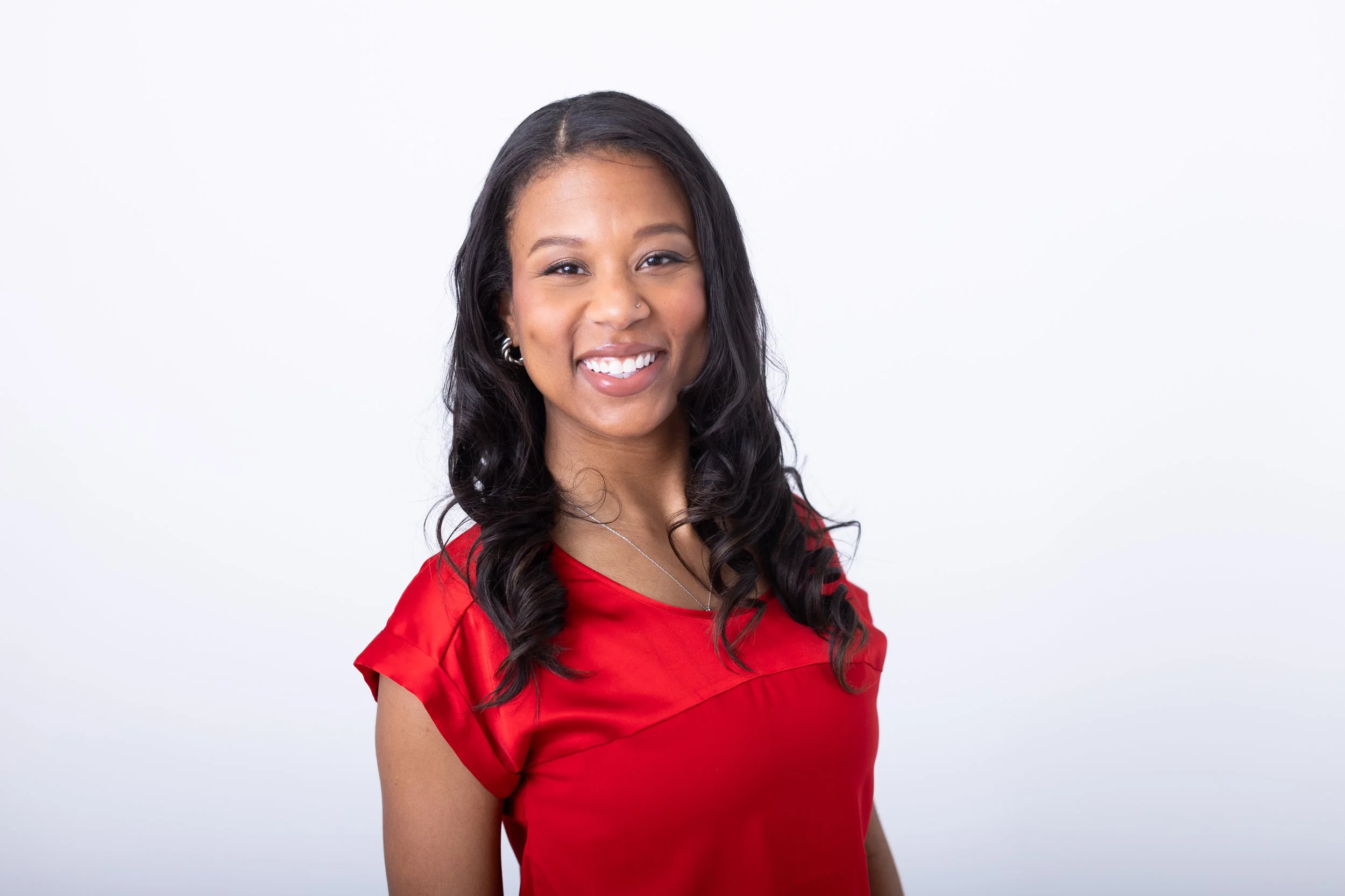 A woman with long wavy hair, wearing a red top, smiling with white teeth, against a plain white background.