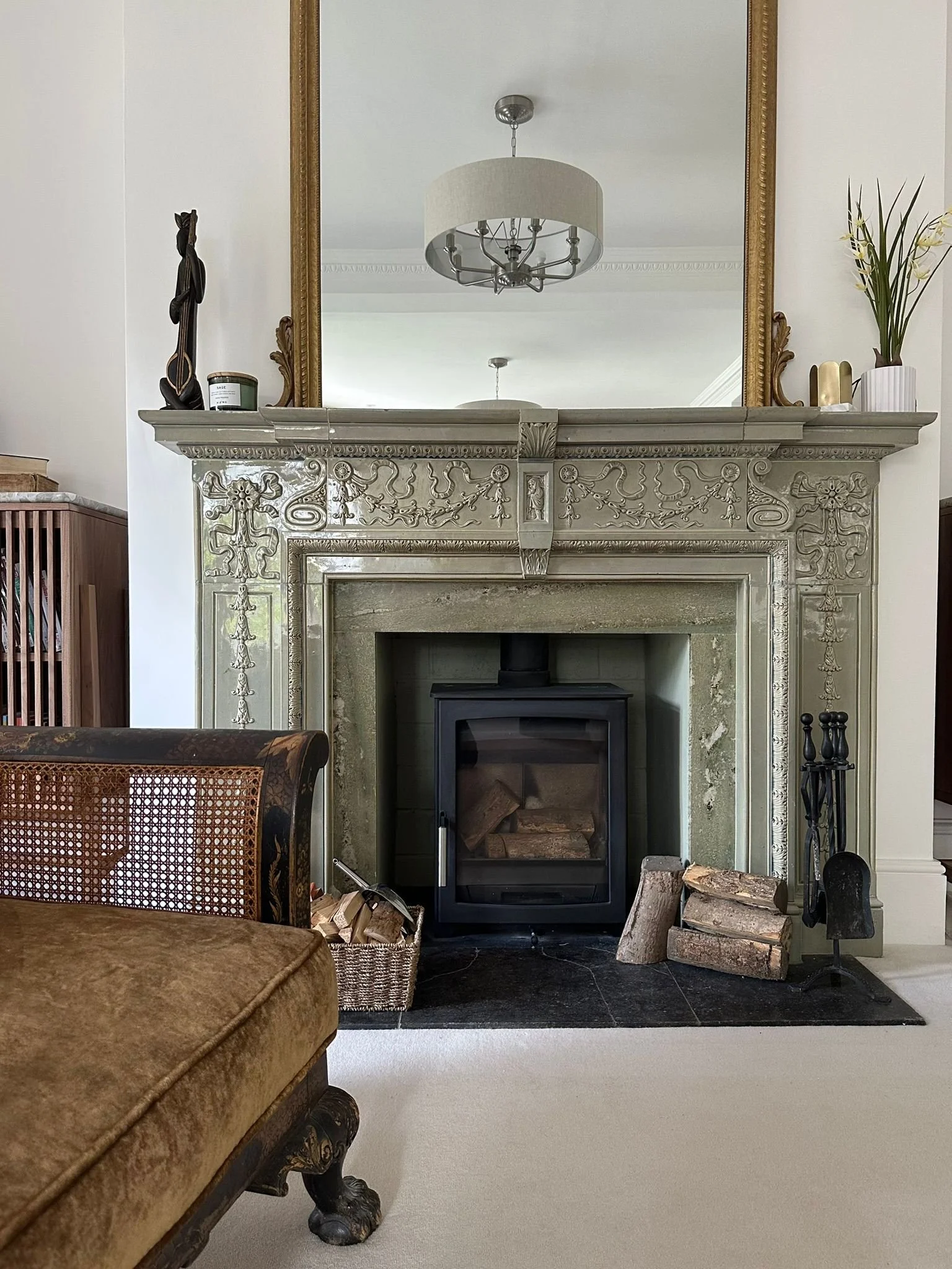 Living room featuring a decorative fireplace with a mirror above it, a chandelier reflected in the mirror, a wooden sculpture on the mantel, a plant in a white striped pot, and logs around a wood stove.