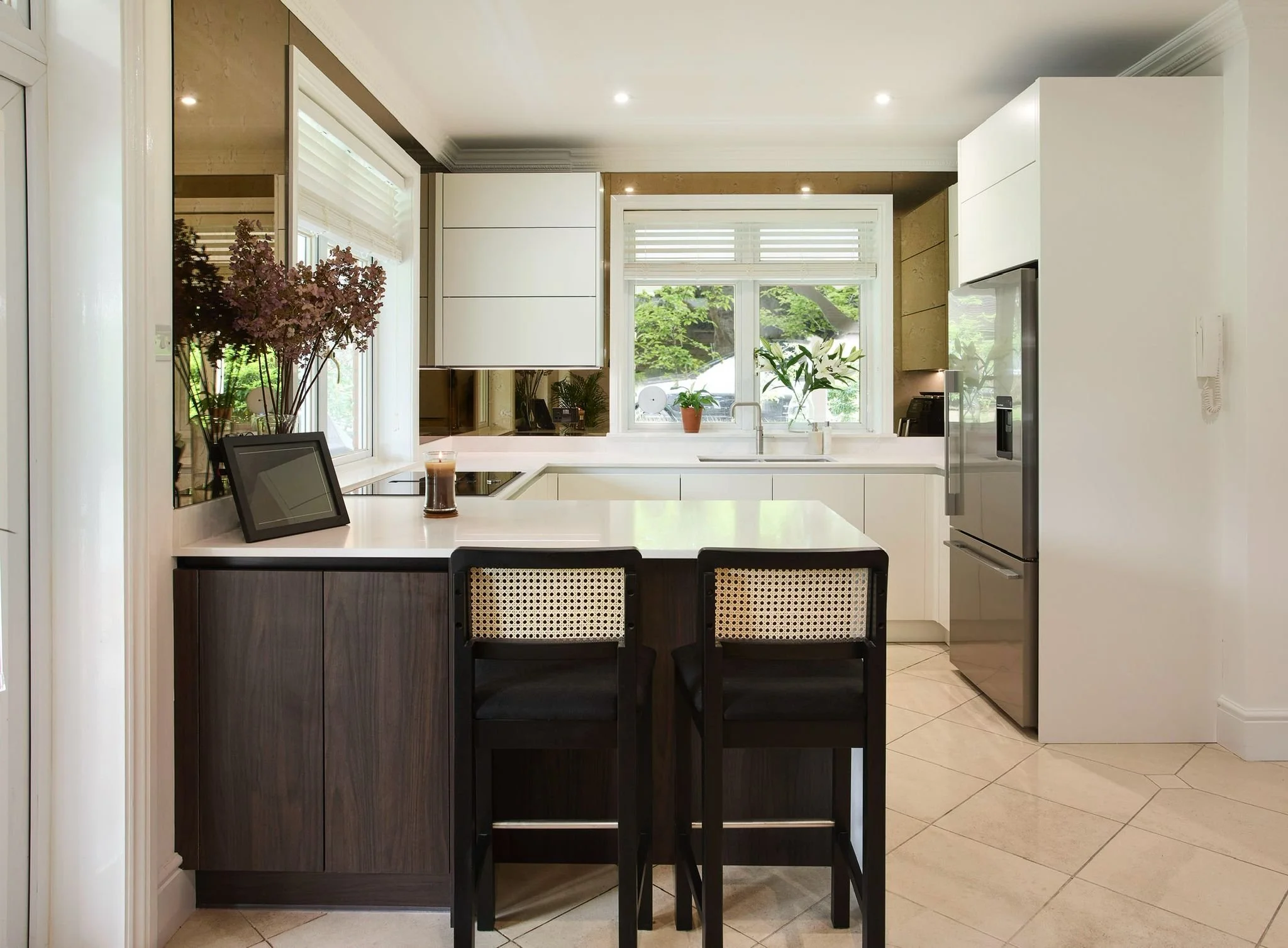 A modern kitchen with white cabinets, a kitchen island with dark wood panels, and a large window with white blinds. There are potted plants on the windowsill, a tablet on the countertop, and a candle. The floor is tiled with beige tiles.