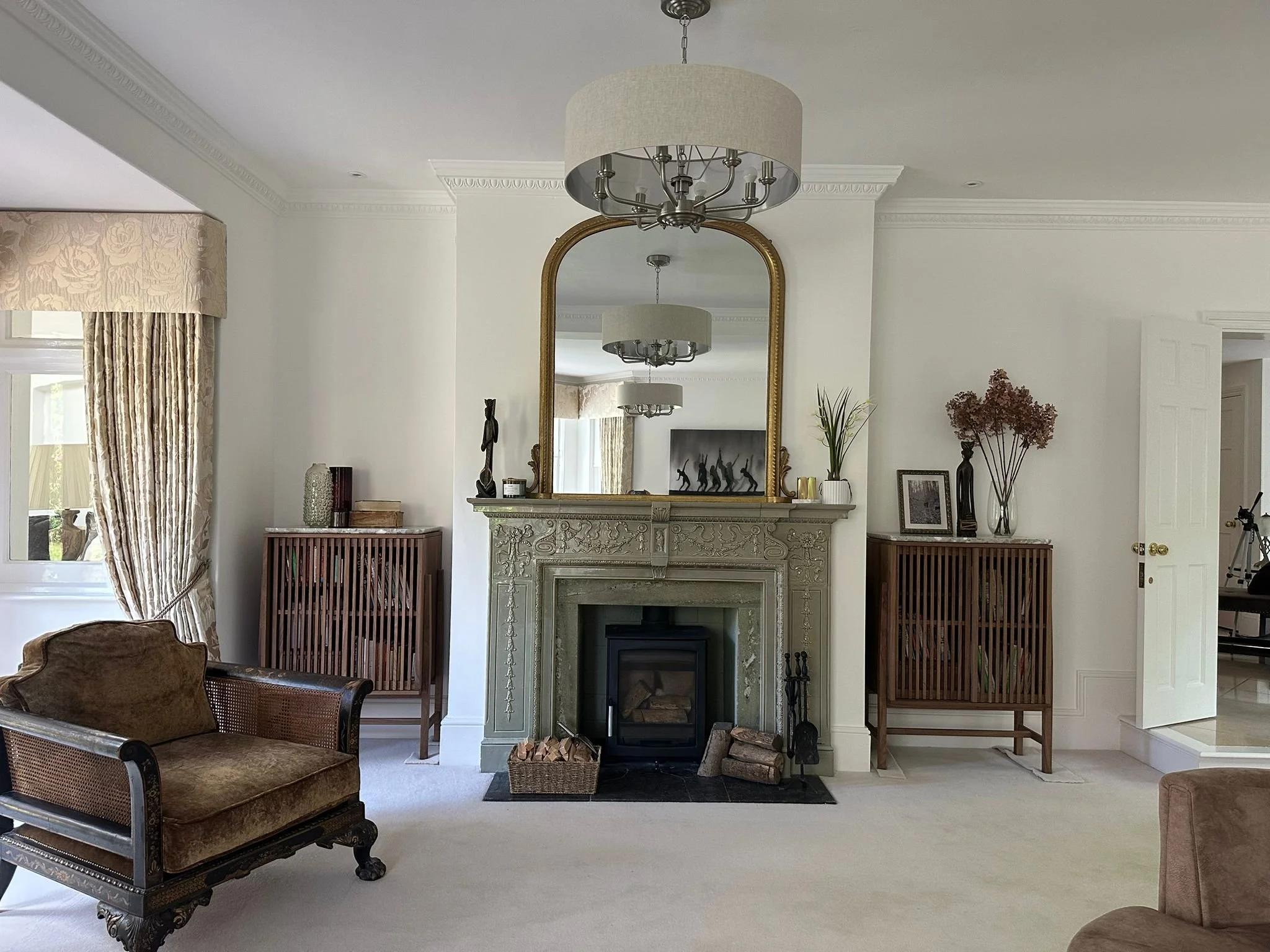 A living room with a fireplace, a large gold-framed mirror above it, and decorative items on the mantel. There is a chandelier hanging from the ceiling and patterned curtains on the window. The room has a light-colored carpet and walls.