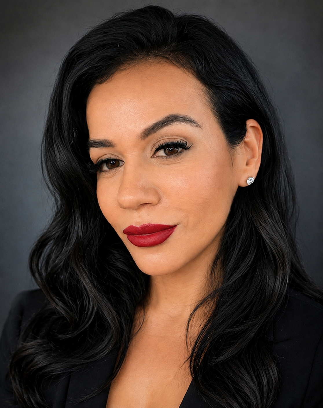 Close-up portrait of a woman with long black wavy hair, red lipstick, and earrings, against a dark gray background.