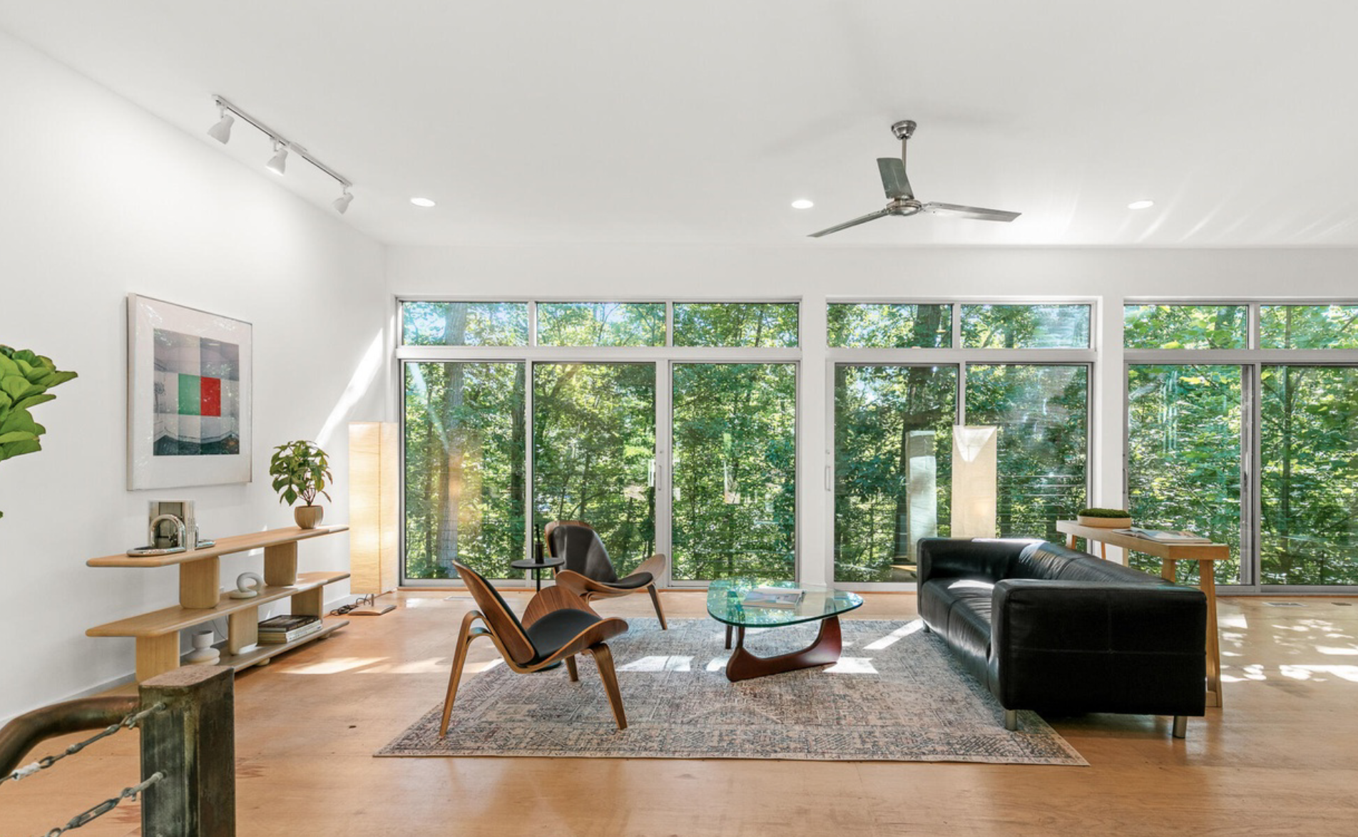 The image shows the exterior of a modern building with large glass windows and doors, surrounded by trees and greenery, with the interior visible through the glass showing furniture and a tall floor lamp.