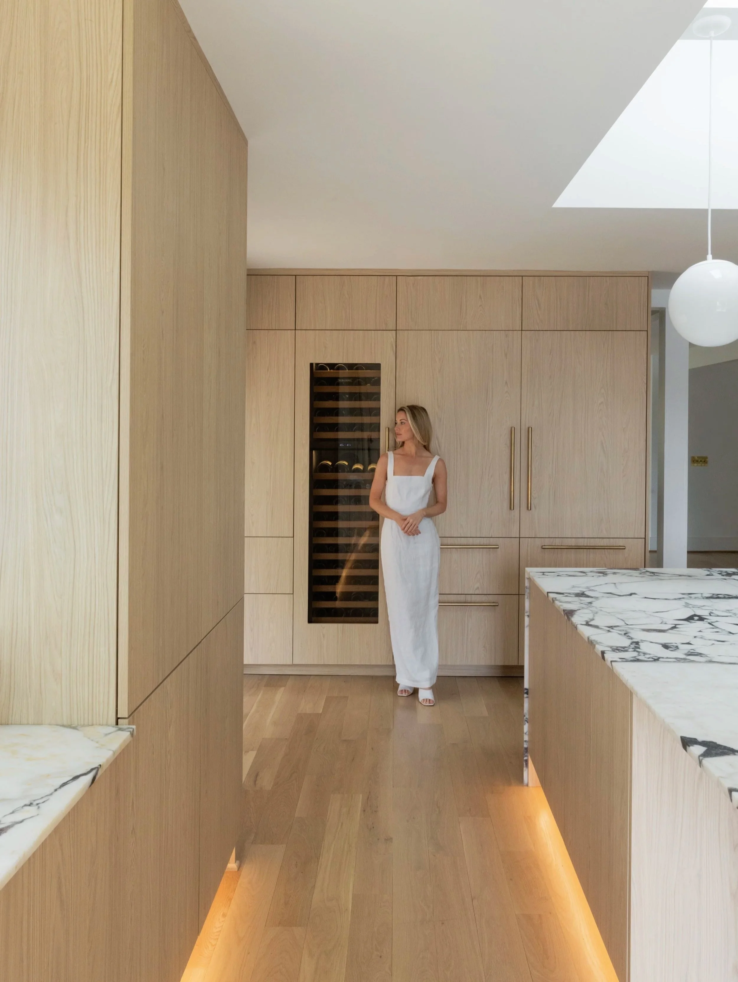 A woman in a white dress standing in a modern kitchen with wood panel cabinets and marble countertops, and a skylight on the ceiling.