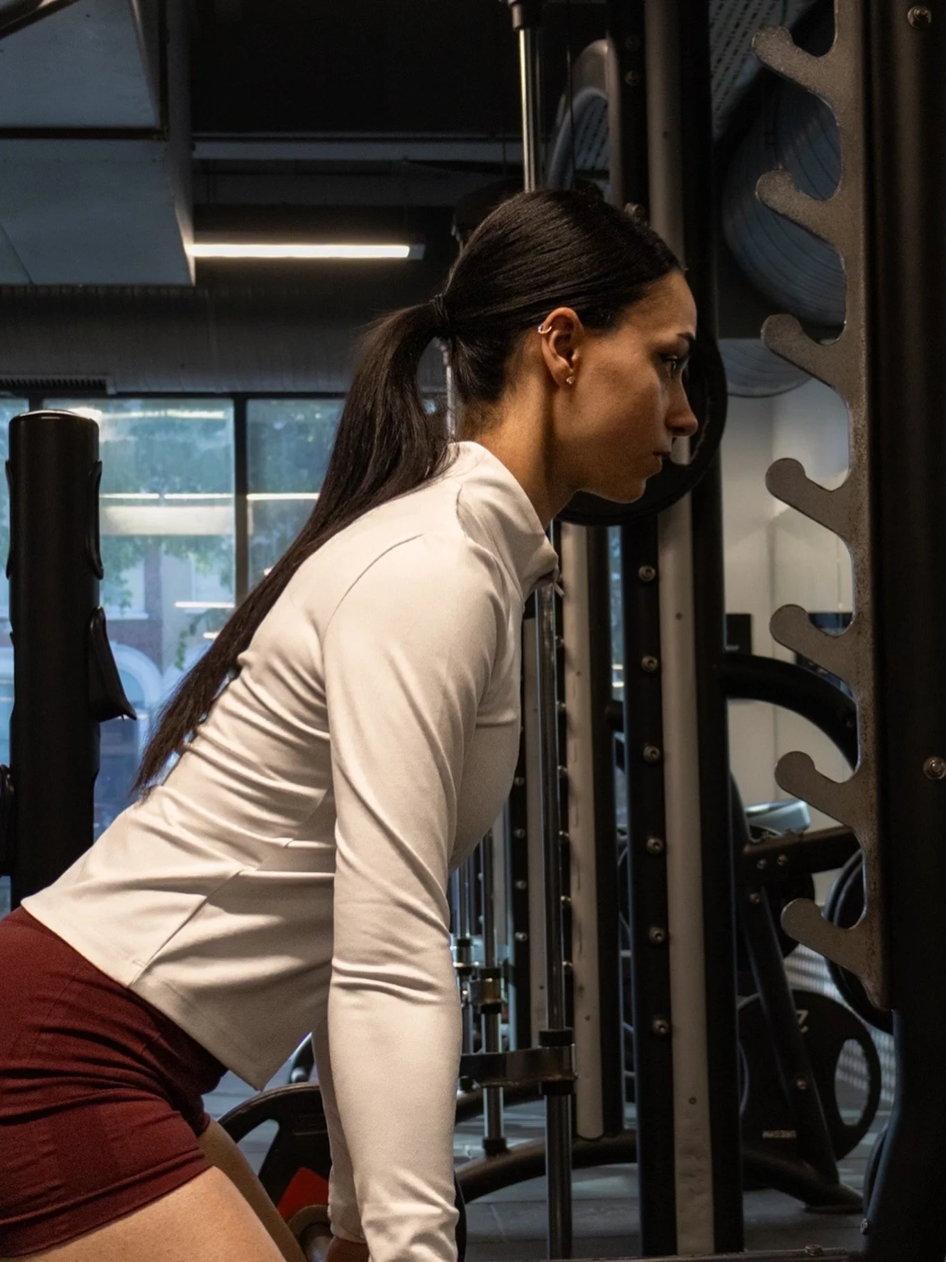 A woman with long dark hair tied back in a ponytail, wearing a beige long-sleeve top and maroon shorts, is working out in a gym. She is leaning forward, possibly doing a bent-over exercise, next to a rack storing black weights.