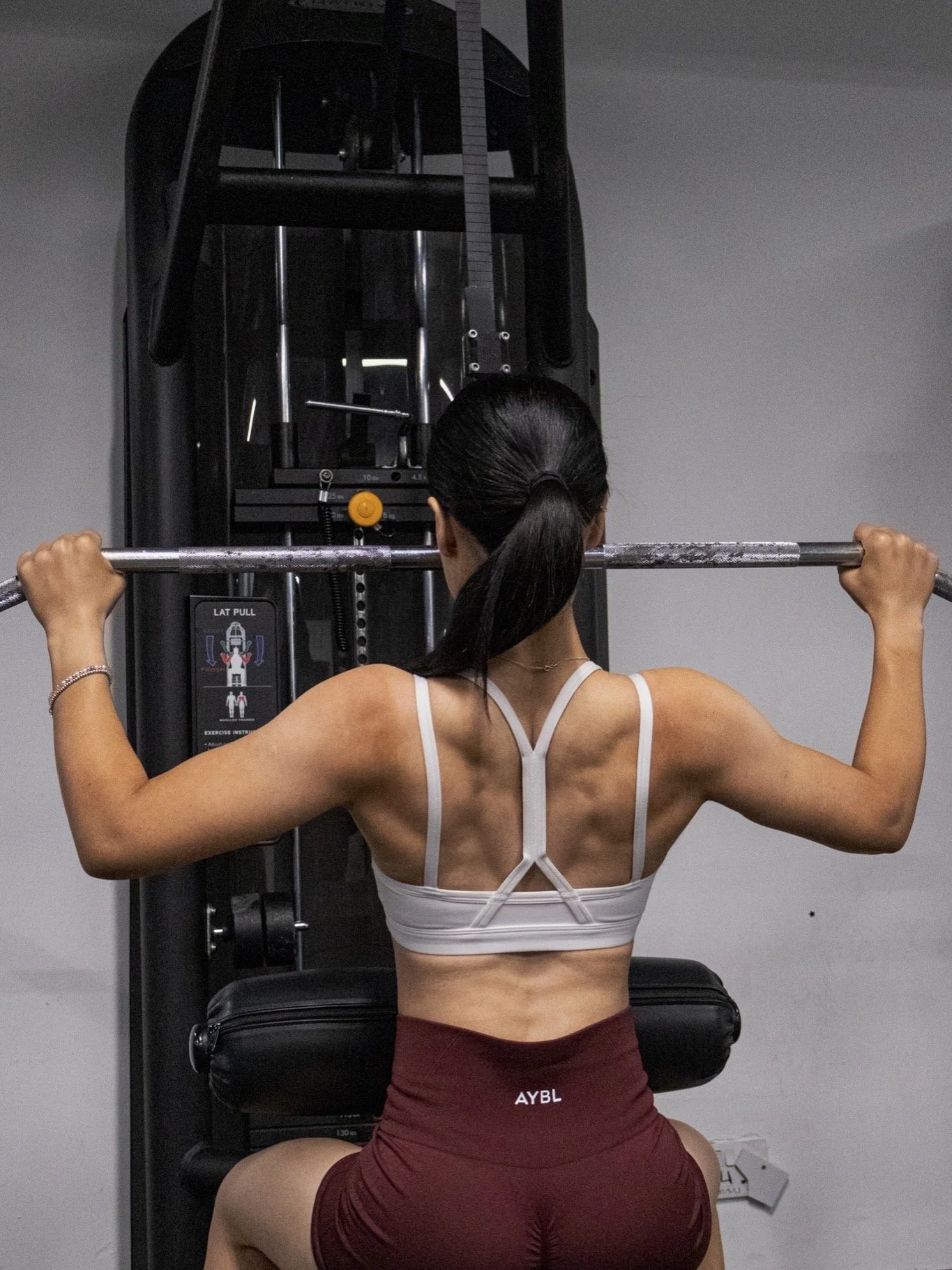 A woman with dark hair tied back, wearing a white sports bra and maroon shorts with 'AYBL' on the waistband, performs a squat using a barbell on a power rack in a gym.
