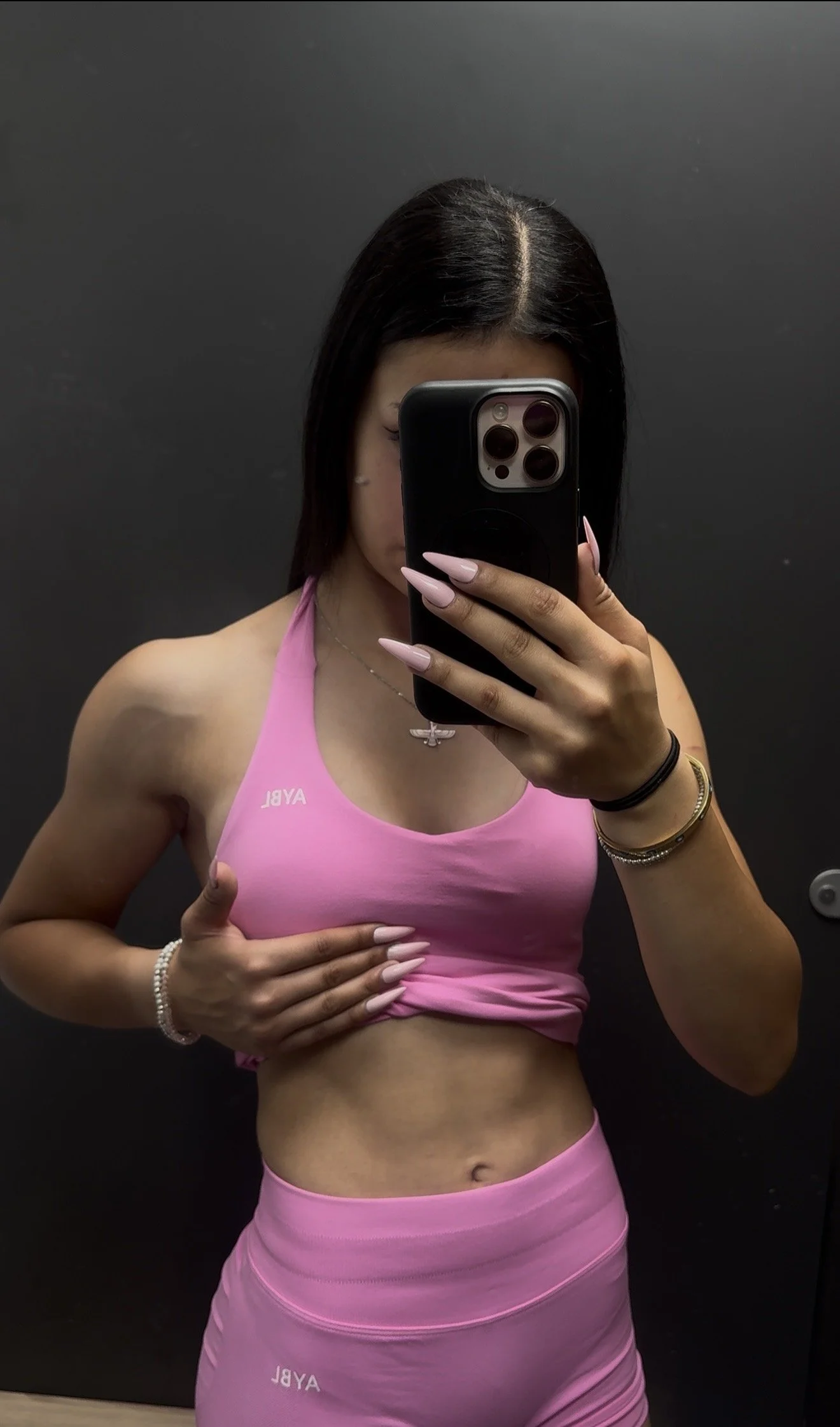 A woman taking a mirror selfie with a smartphone in an elevator. She is wearing a pink athletic top and matching pink leggings, with a silver necklace, bracelets, and long pink nails. She is covering her face with the phone, showing part of her dark hair and toned physique.