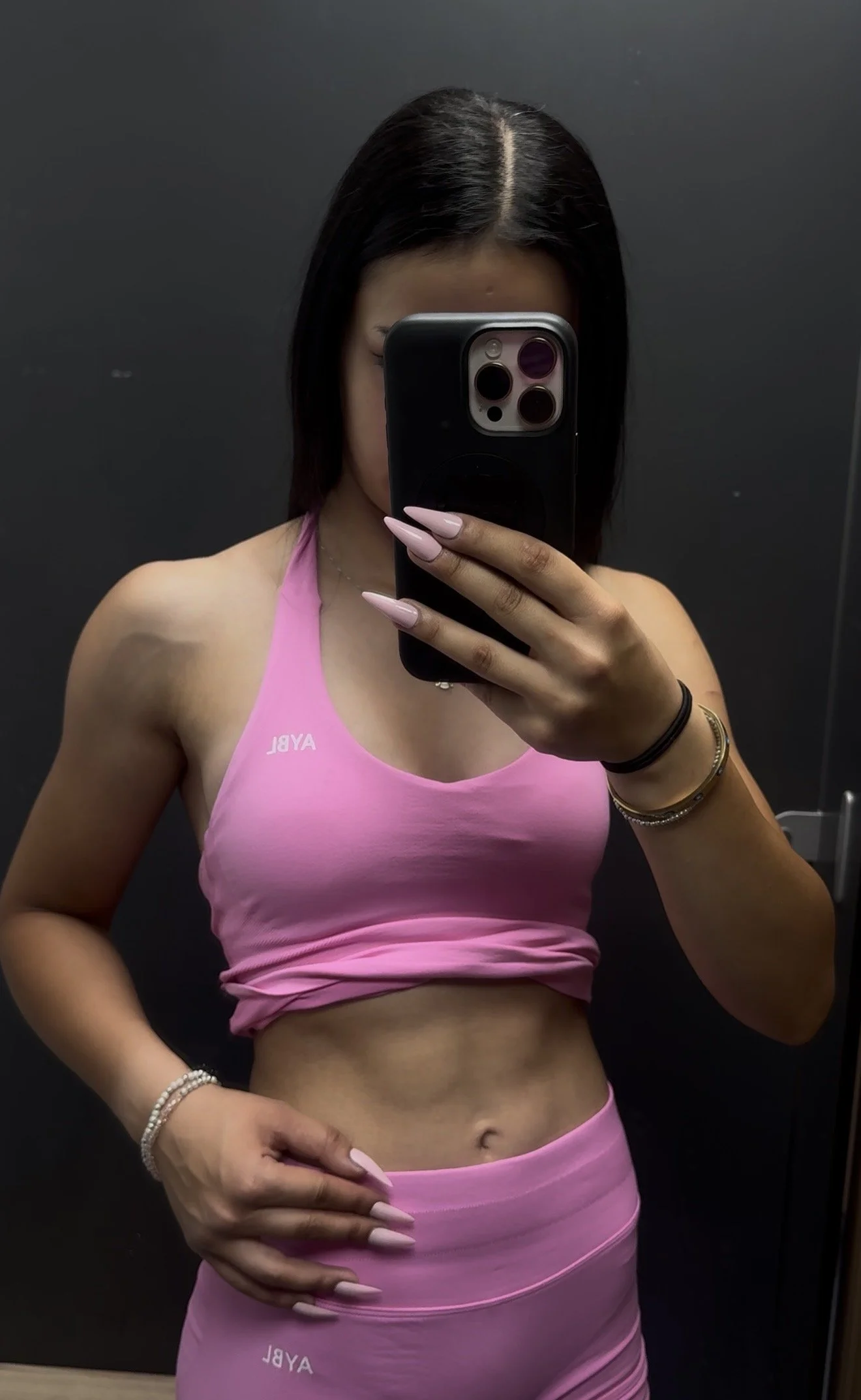 A woman taking a mirror selfie in a dark room or elevator, wearing a pink sports bra with the word 'AYBL' on it and matching pink leggings, showing her toned stomach.
