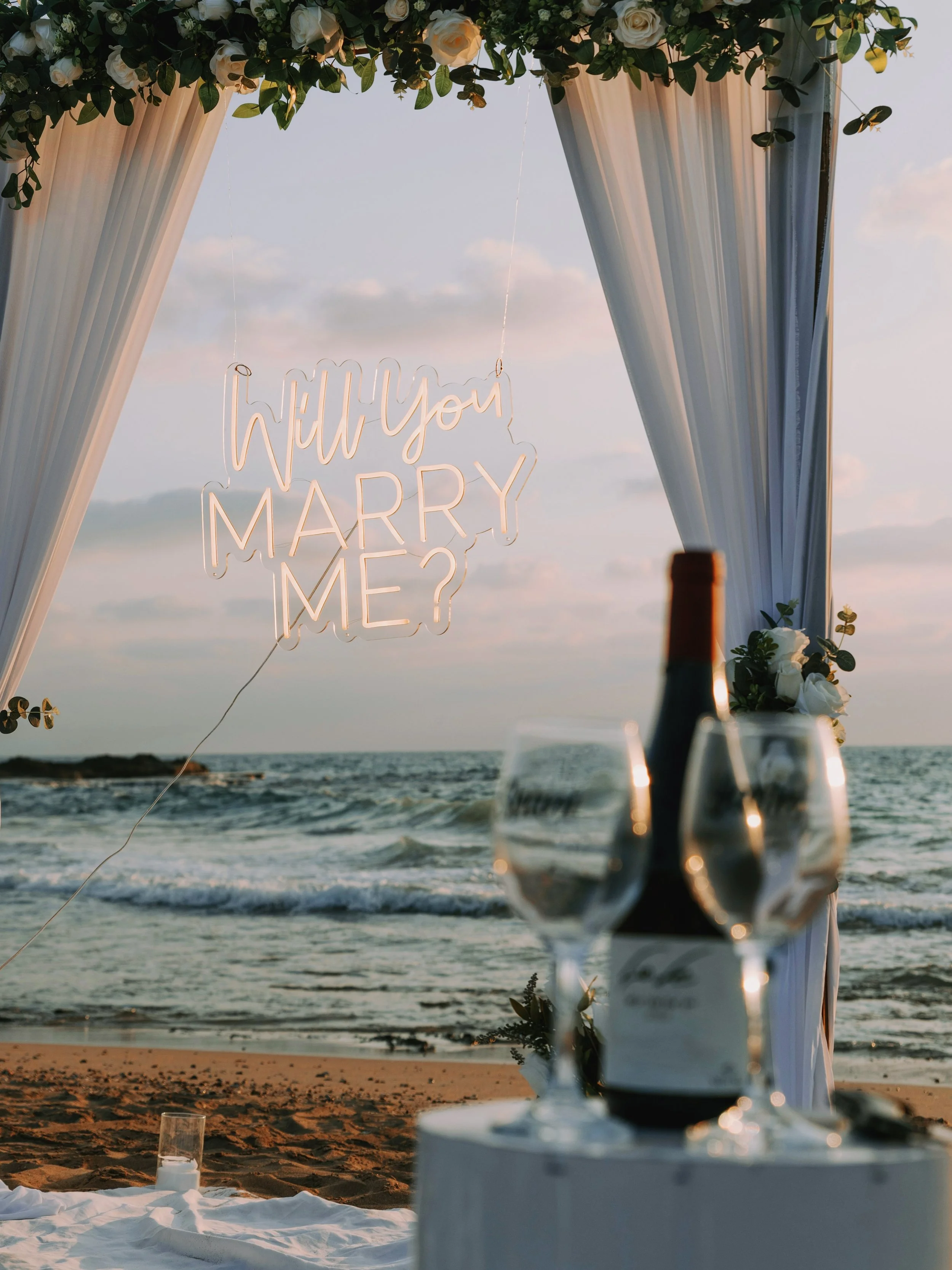 A beach wedding setup with white curtains, floral decorations, and a sign that reads 'Will you marry me?' hanging from the top. In the foreground, there are two glasses of champagne and a bottle on a table, with the ocean and a sunset in the background.