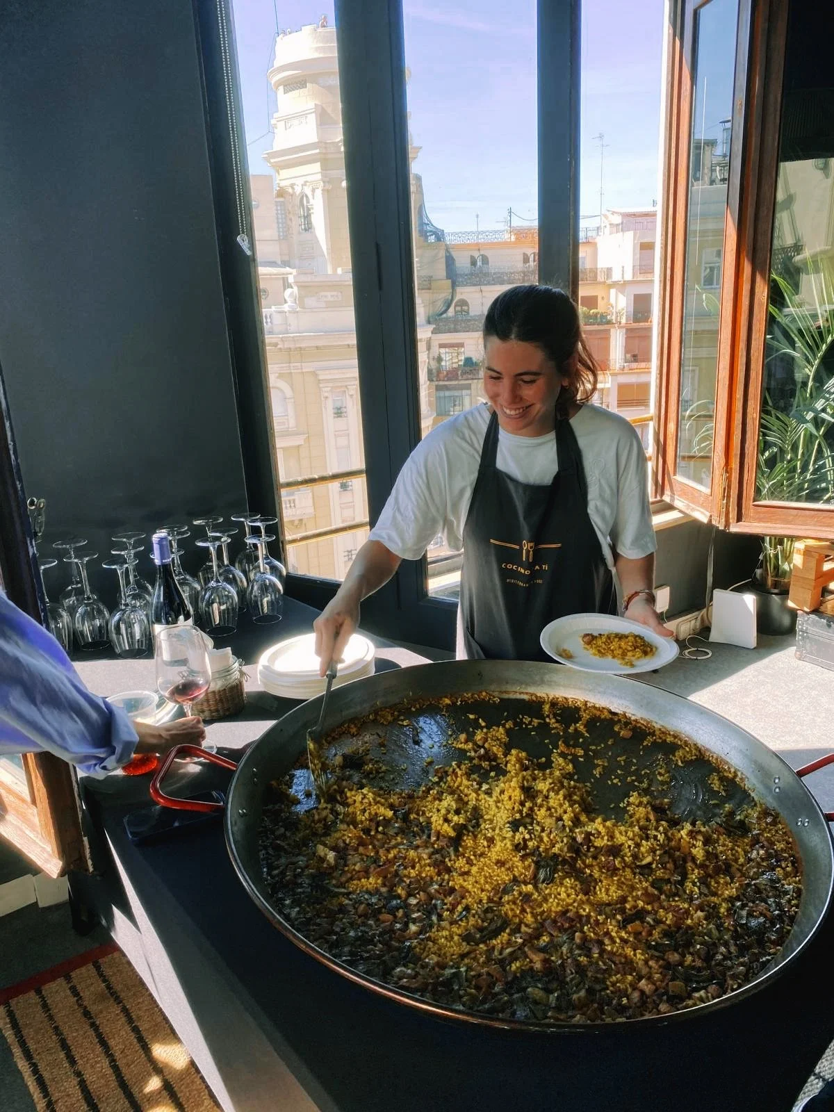 A woman serving food from a large skillet of yellow and brown rice, standing indoors near a window with a cityscape in the background. She is smiling and holding a plate, wearing a black apron over a white T-shirt.