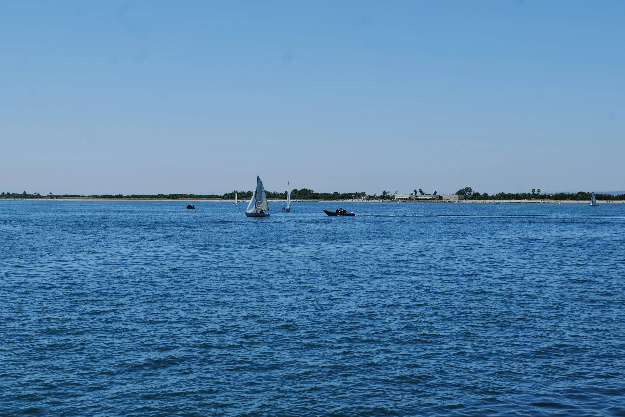 A scenic view of a body of water with several sailboats and small boats, a distant shoreline with trees and buildings, under a clear blue sky.
