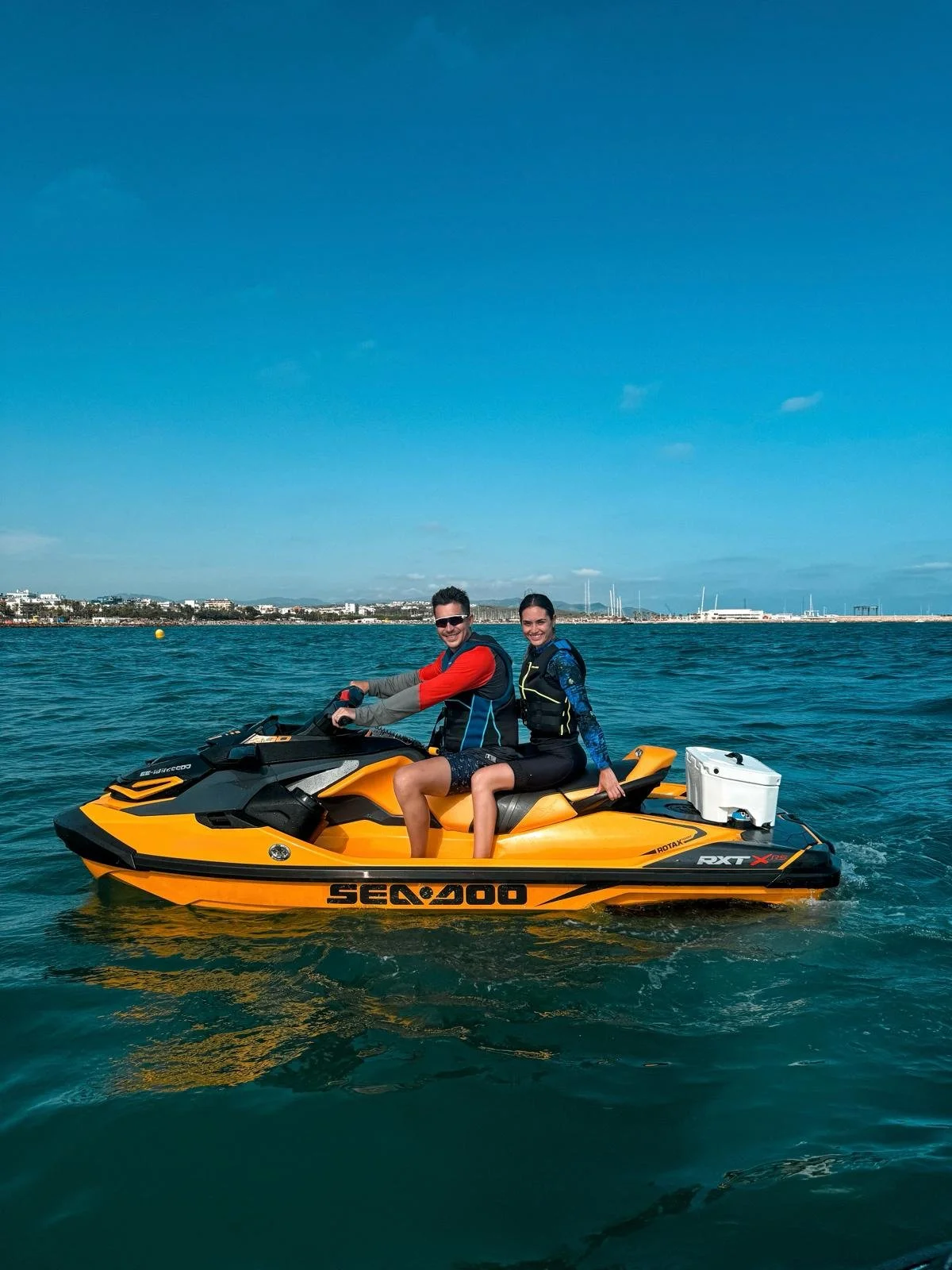 Two people riding a yellow and black Sea-Doo jet ski on the water, with a shoreline and buildings in the background under a blue sky.