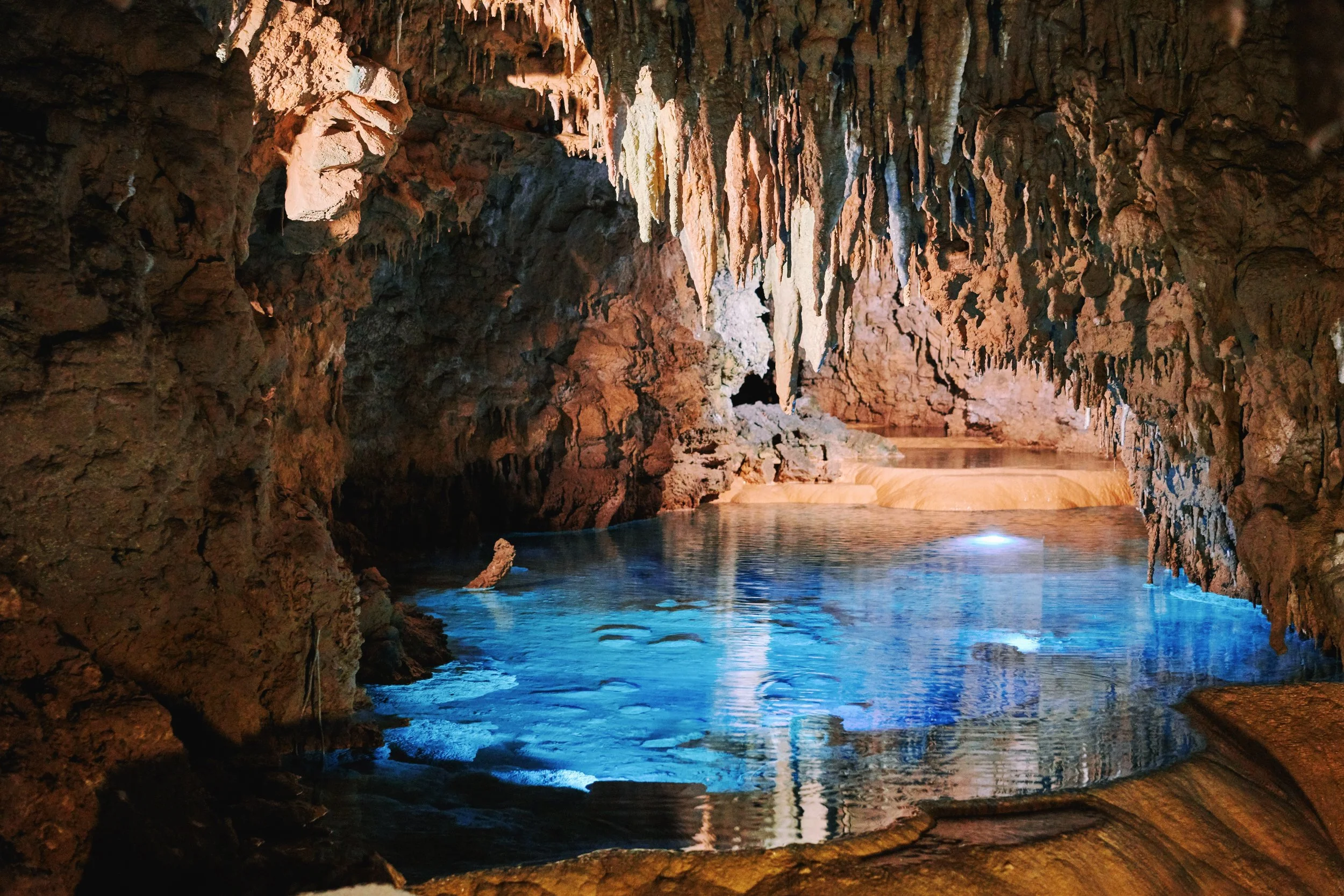 Inside a cave with stalactites hanging from the ceiling and a pool of water reflecting the cave walls.
