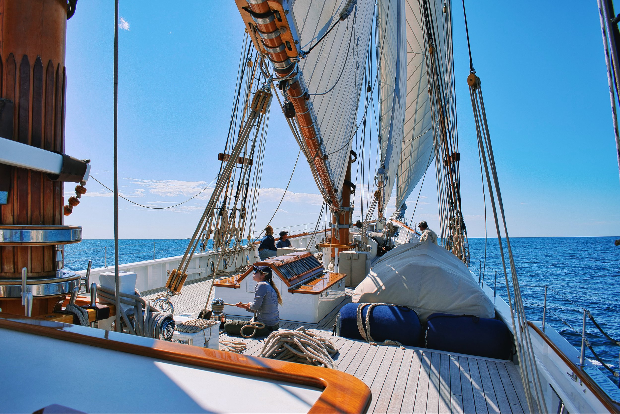 View from a sailing ship deck with people working, blue sky, ocean, and ship's rigging.
